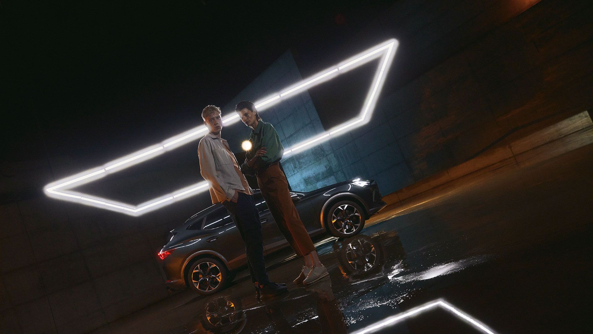 Dva osebi stojita pred sodobnim avtomobilom CUPRA Umbrella, ki je osvetljen z geometrijsko oblikovano LED lučjo. Površina tal je mokra in ustvarja odsev luči ter avtomobila.