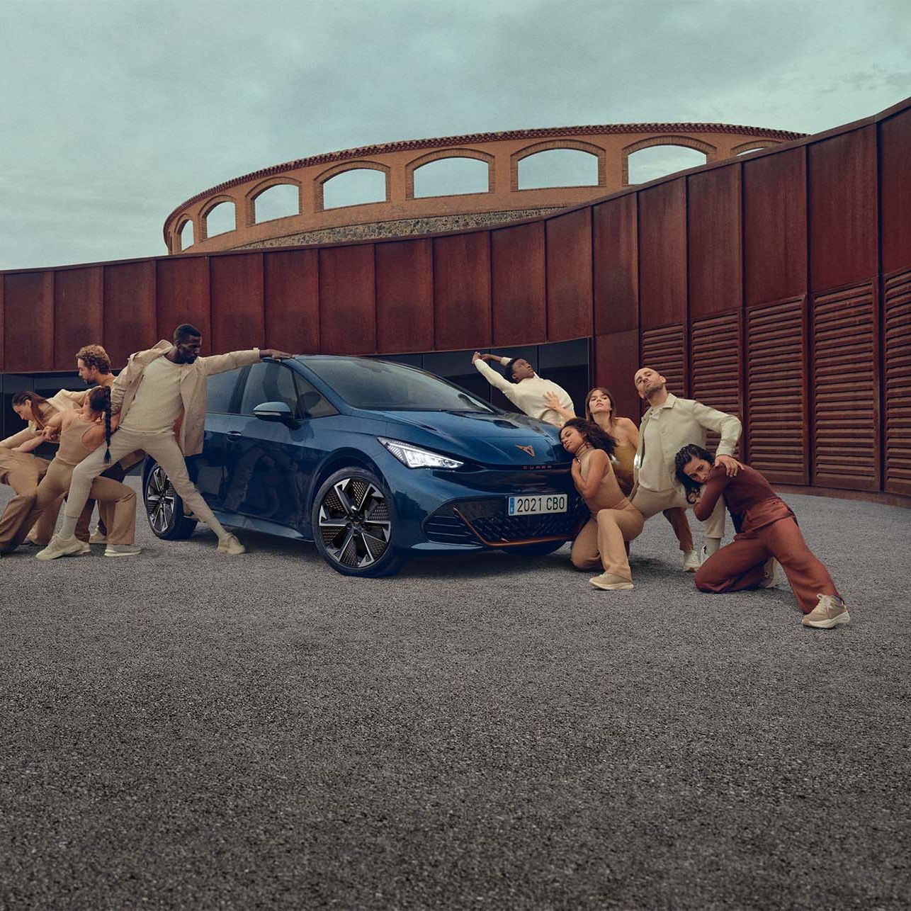 Več ljudi v različnih plesnih pozah, oblečenih v nevtralne barve, obkrožajo temno moder športni avtomobil Cupra Born, postavljen na utrjenem dvorišču z modernimi lesenimi stavbami v ozadju.