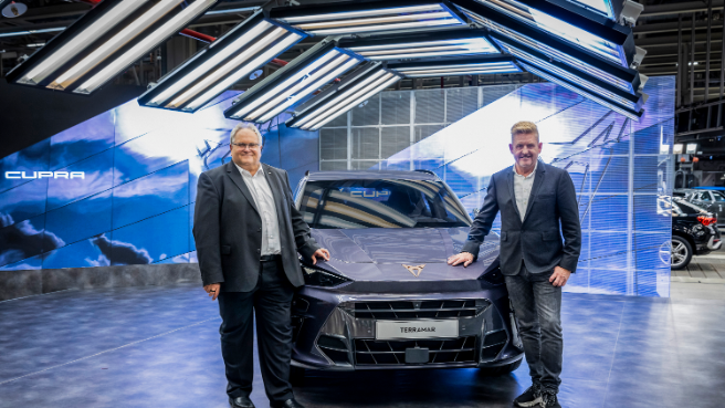 Dva moška stojita pred avtomobilom CUPRA Terramar na avtomobilskem sejmu. Ozadje je osvetljeno z več lučmi, viden je velik zaslon. Avtomobili so razstavljeni v ozadju znotraj razstavnega prostora.