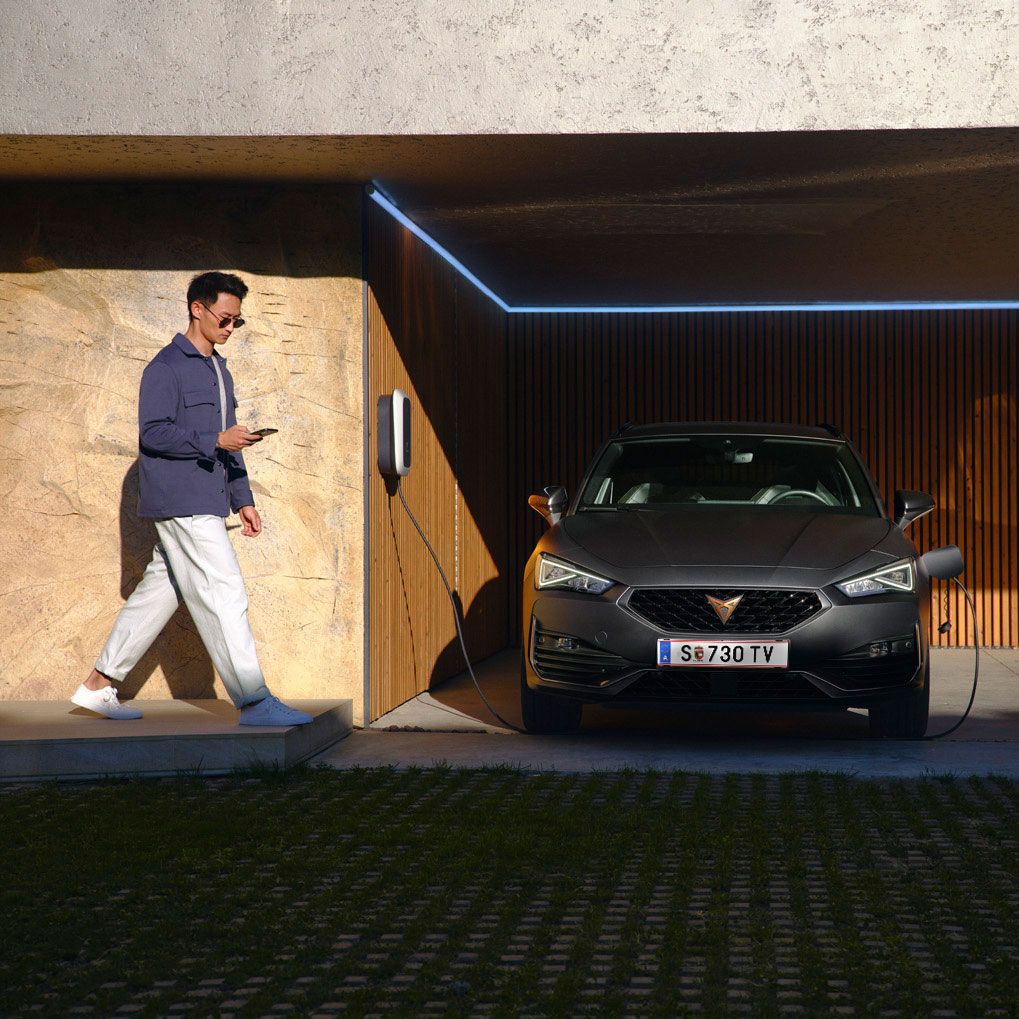 Moški hodi mimo parkiranega električnega avtomobila CUPRA Leon SP, ki se polni v garaži sodobnega videza.