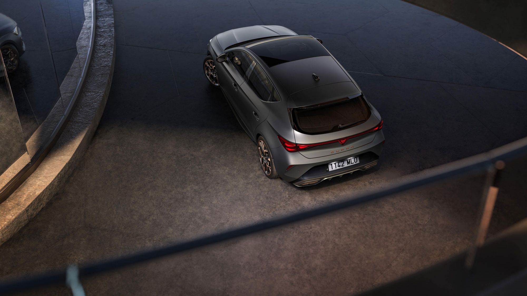 Sodoben športni avto Cupra Leon, parkiran na ukrivljenem in dvignjenem parkirišču. Avto je sive barve z elegantnim dizajnom. Okolje je moderno, z betonskimi in steklenimi elementi.