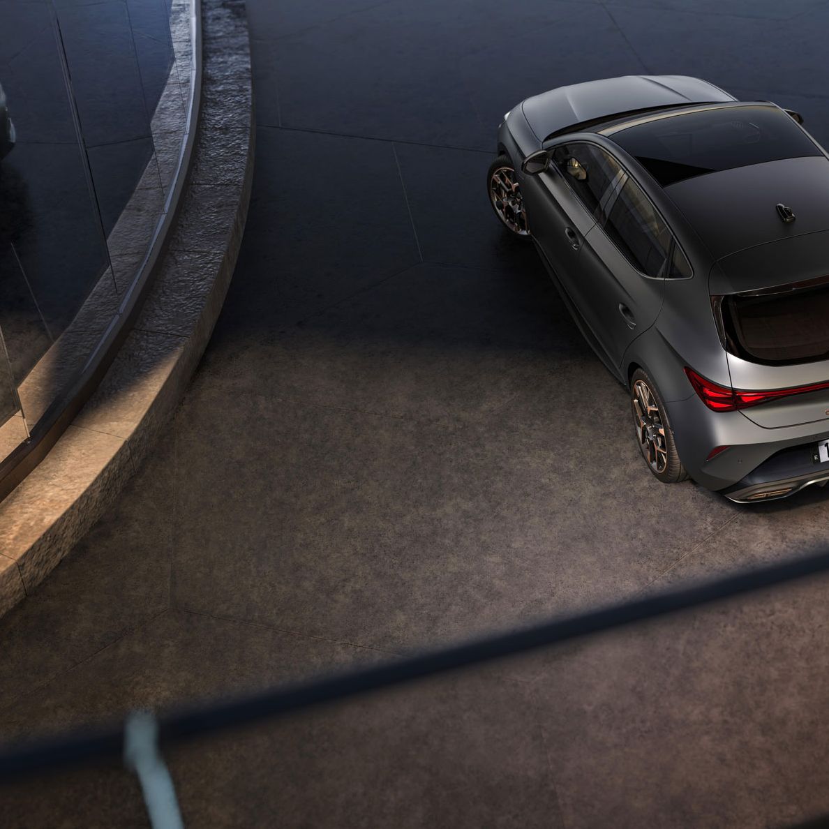 Sodoben športni avto Cupra Leon, parkiran na ukrivljenem in dvignjenem parkirišču. Avto je sive barve z elegantnim dizajnom. Okolje je moderno, z betonskimi in steklenimi elementi.