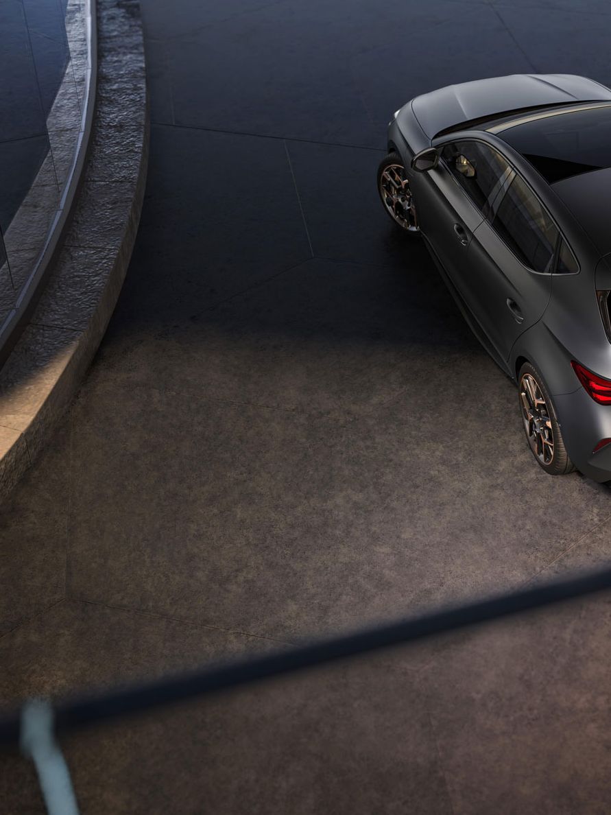 Sodoben športni avto Cupra Leon, parkiran na ukrivljenem in dvignjenem parkirišču. Avto je sive barve z elegantnim dizajnom. Okolje je moderno, z betonskimi in steklenimi elementi.