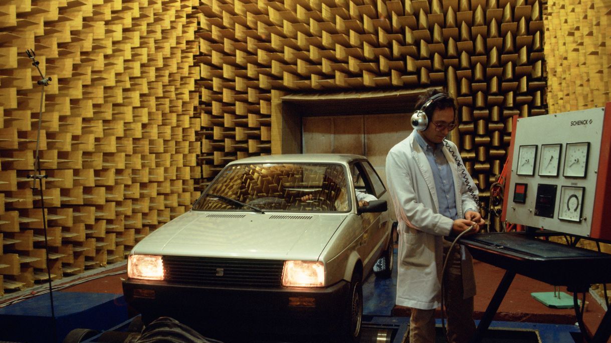 Avtomobil znamke SEAT z vključenimi žarometi stoji v zvočno izolirani komori z akustičnimi penastimi paneli, medtem ko tehnik v belem plašču in s slušalkami upravlja merilno napravo ob testni mizi. Slika je stara približno 50 let.