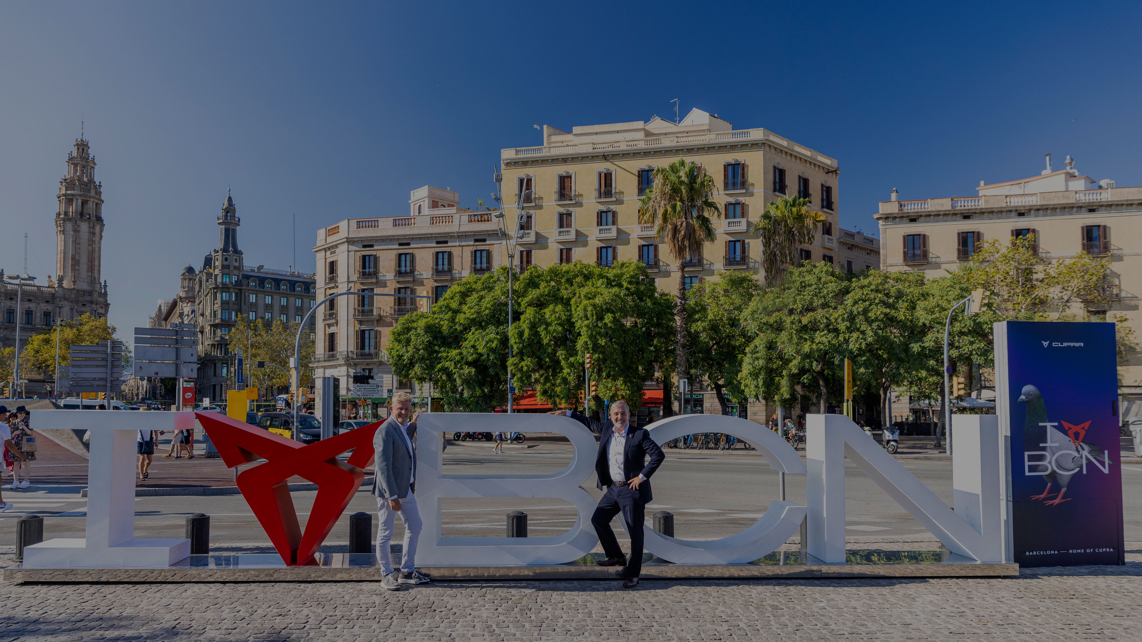 Dva moška stojita na mestnem trgu v Barceloni, ob veliki napisni skulpturi »I love BCN«, pri čemer je simbol za »love« oblikovan iz logotipa znamke CUPRA. V ozadju so zgodovinske stavbe, palme in živahno mestno dogajanje v sončnem dnevu.