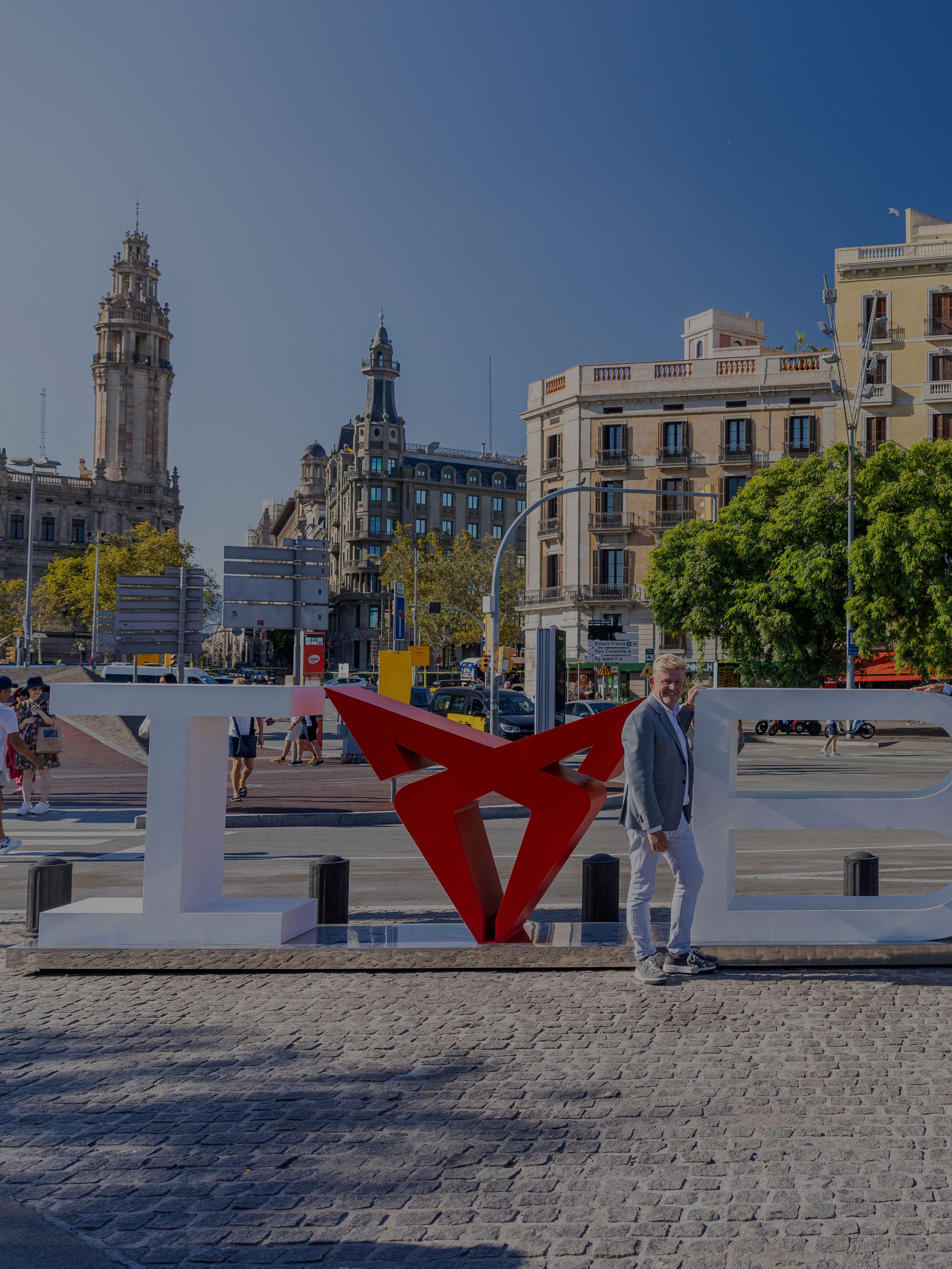 Dva moška stojita na mestnem trgu v Barceloni, ob veliki napisni skulpturi »I love BCN«, pri čemer je simbol za »love« oblikovan iz logotipa znamke CUPRA. V ozadju so zgodovinske stavbe, palme in živahno mestno dogajanje v sončnem dnevu.