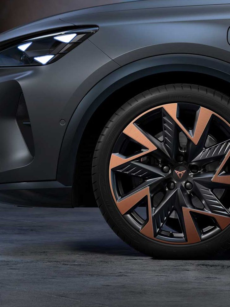 Sprednji del mat temno sivega vozila CUPRA Formentor s poudarkom na sprednjem levem kolesu in sodobno oblikovanih črno-oranžnih platiščih. V ozadju je zamegljeno mestno okolje ob mraku.