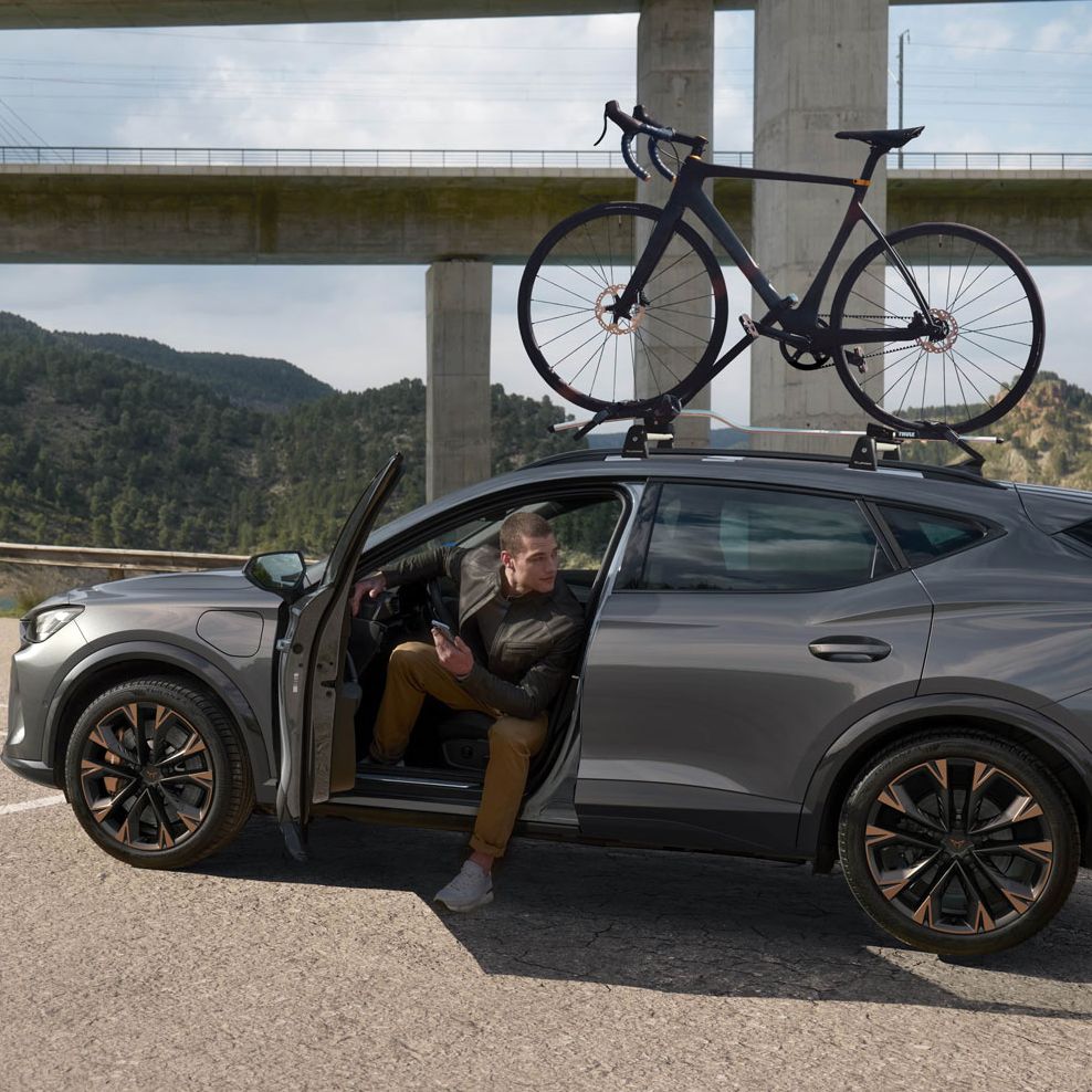 Voznik sedi na sedežu avtomobila CUPRA Formentor PA, parkiranega ob cesti. Na strehi avtomobila je pritrjeno kolo. V ozadju so vidni viadukt in naravna pokrajina.