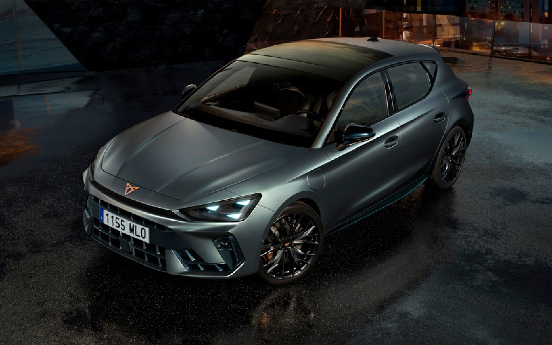 CUPRA Leon metal sive barve, parkiran na mokrem, osvetljenem urbanem območju ponoči. Avto ima ostre, aerodinamične linije.