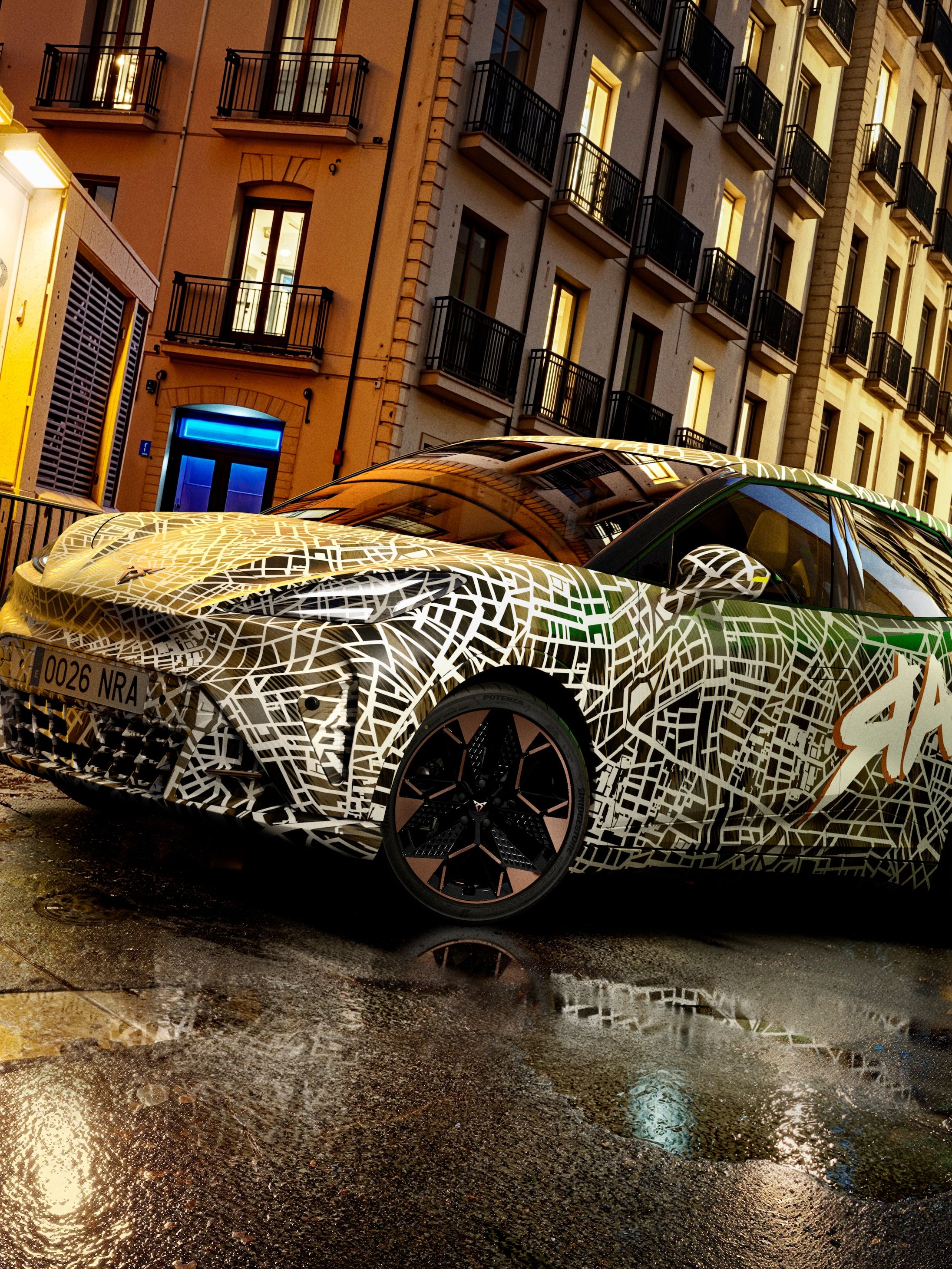 Avtomobil CUPRA Raval z grafiti vzorcem parkiran na mokri ulici mestne četrti med stanovanjskimi blokovskimi hišami, osvetljen z ulicnimi lucmi, v ozadju pa velik grafit na steni.