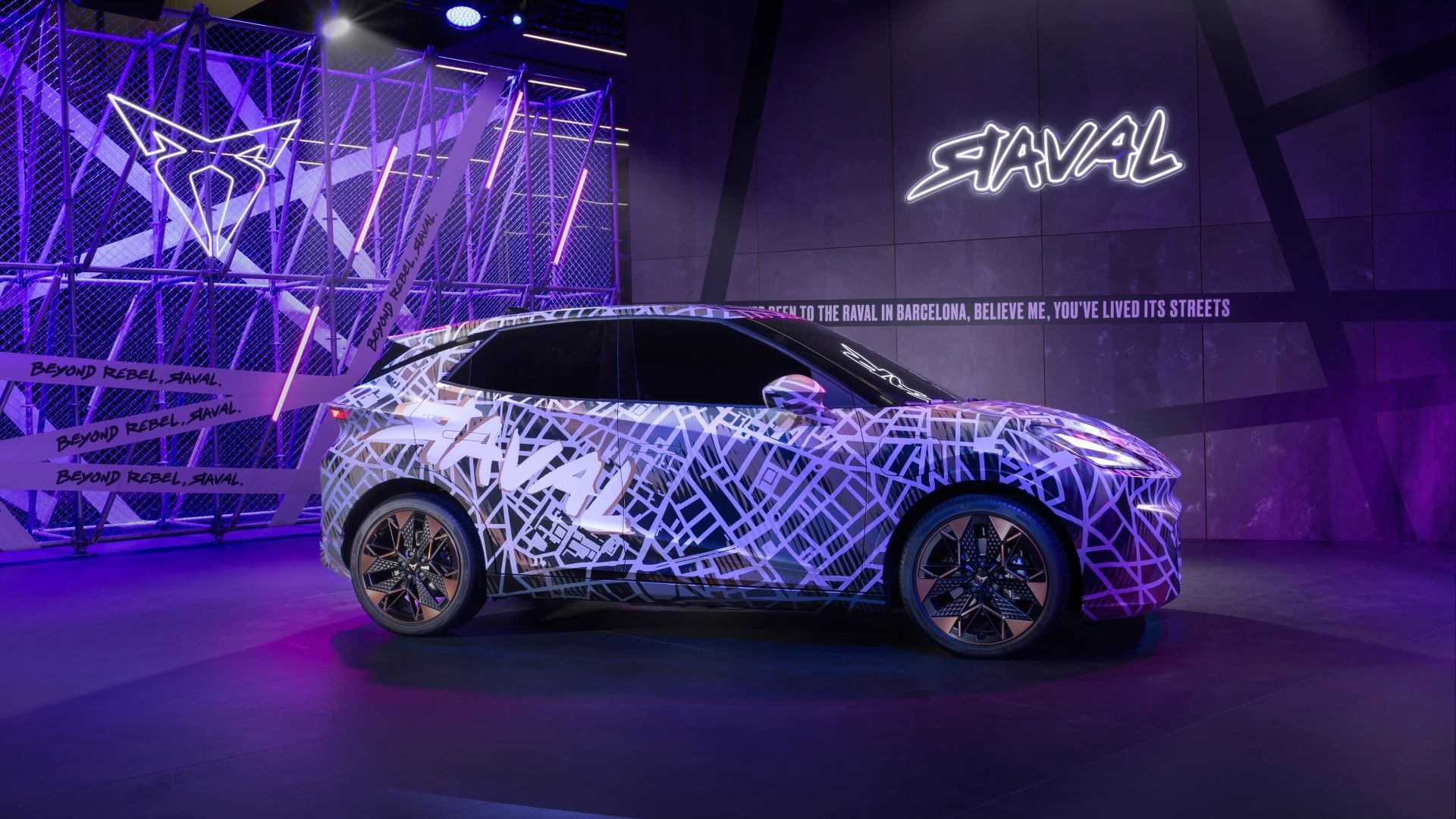 CUPRA Raval Dynamic je razstavljen v prostoru z vijoličnimi in modrimi osvetlitvami. Vozilo ima moderno geometrično poslikavo z vzorci, ki spominjajo na mestne ulice. Ozadje vključuje velik grafični element in napise.