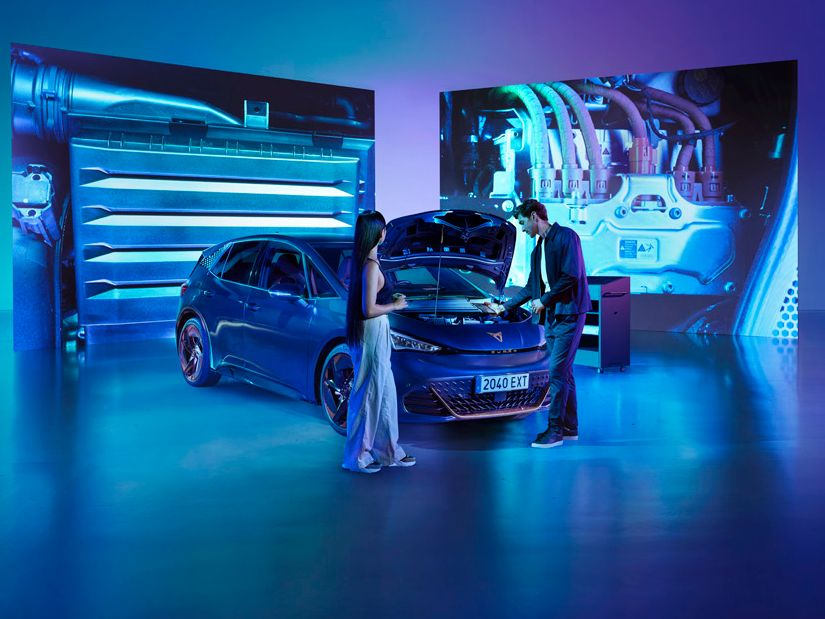 Ženska in moški stojita v studiu ob modrem avtomobilu CUPRA Born z odprtim pokrovom motorja v svetlo modri osvetlitvi. Za avtomobilom so veliki zasloni, ki prikazujejo dele motorja.