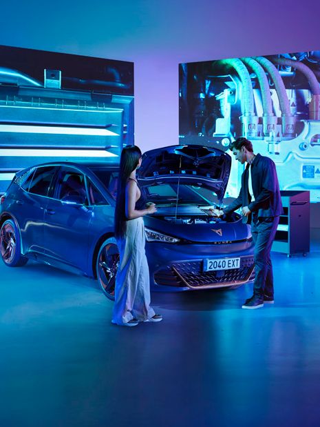 Ženska in moški stojita v studiu ob modrem avtomobilu CUPRA Born z odprtim pokrovom motorja v svetlo modri osvetlitvi. Za avtomobilom so veliki zasloni, ki prikazujejo dele motorja.