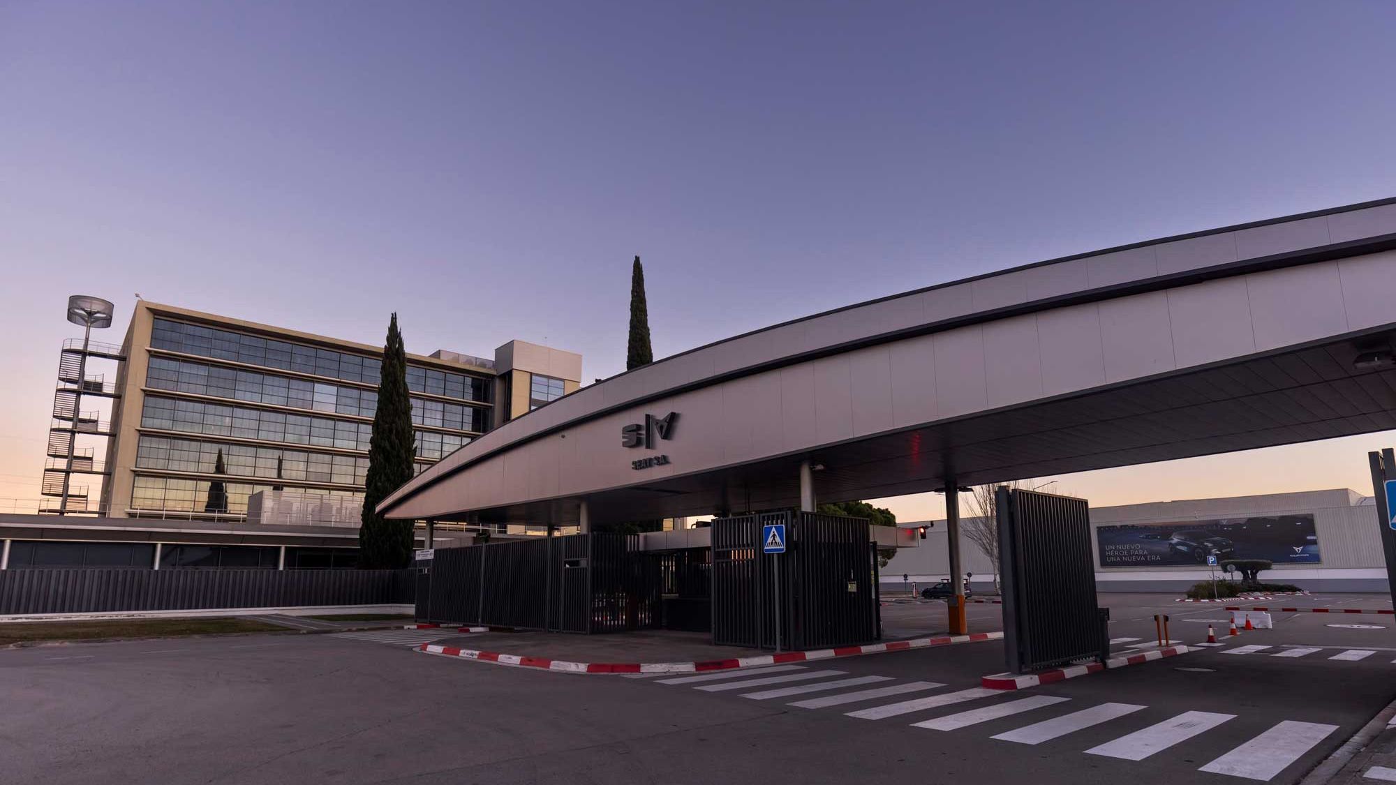 Poslovna stavba z modernim steklenim zunanjim izgledom ob sončnem zahodu. Pred stavbo je nadstrešek in označene prehode za pešce. 