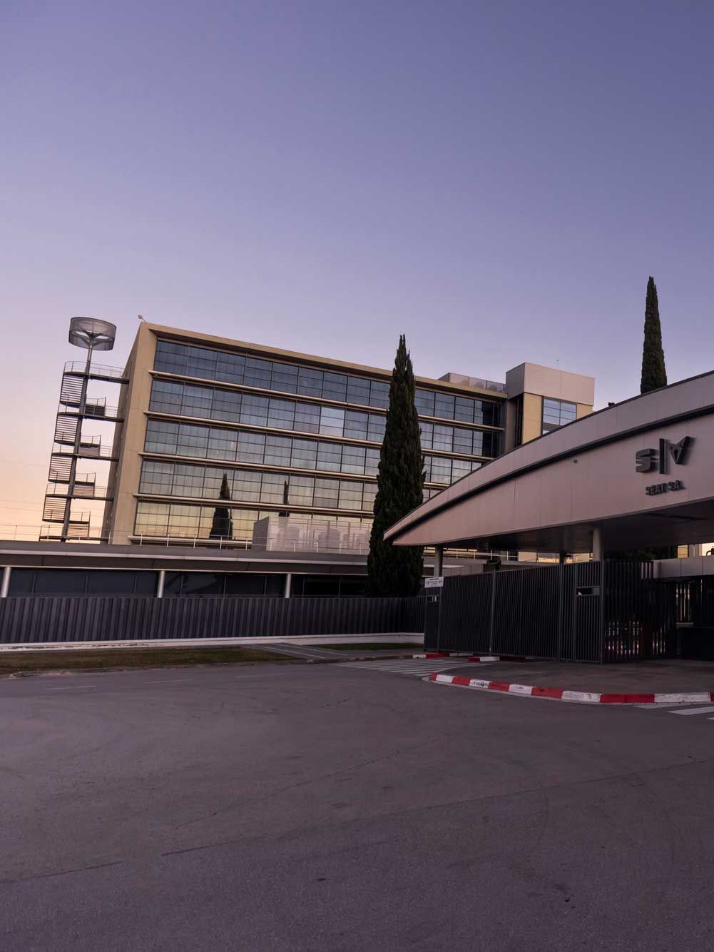 Poslovna stavba z modernim steklenim zunanjim izgledom ob sončnem zahodu. Pred stavbo je nadstrešek in označene prehode za pešce. 