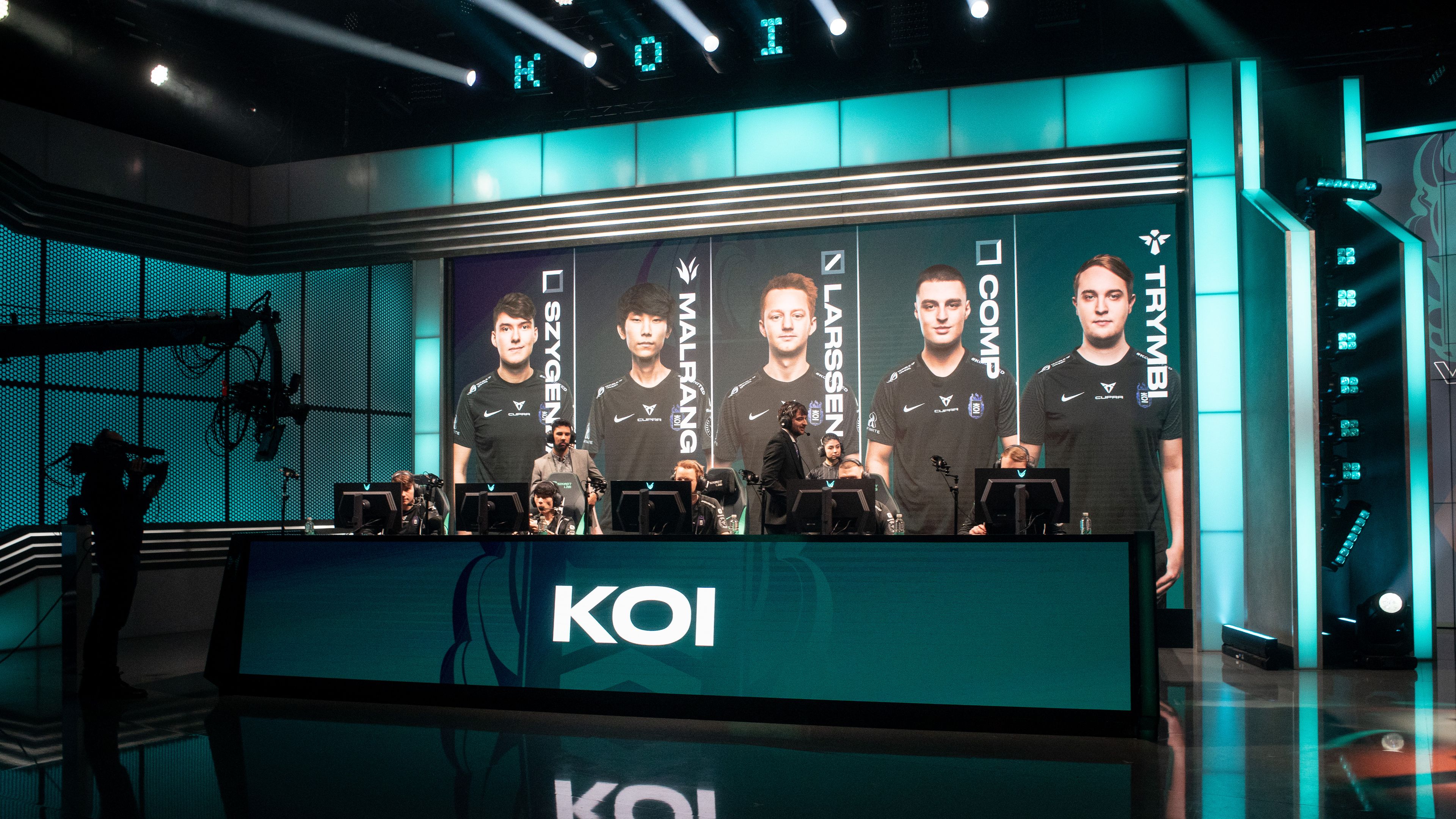 Na odru je ekipa KOI, sestavljena iz petih članov. V ozadju so vidne osvetljene velike zaslone z njihovimi imeni in obrazi. Ekipa sedi za računalniki, pripravljena na tekmovanje.