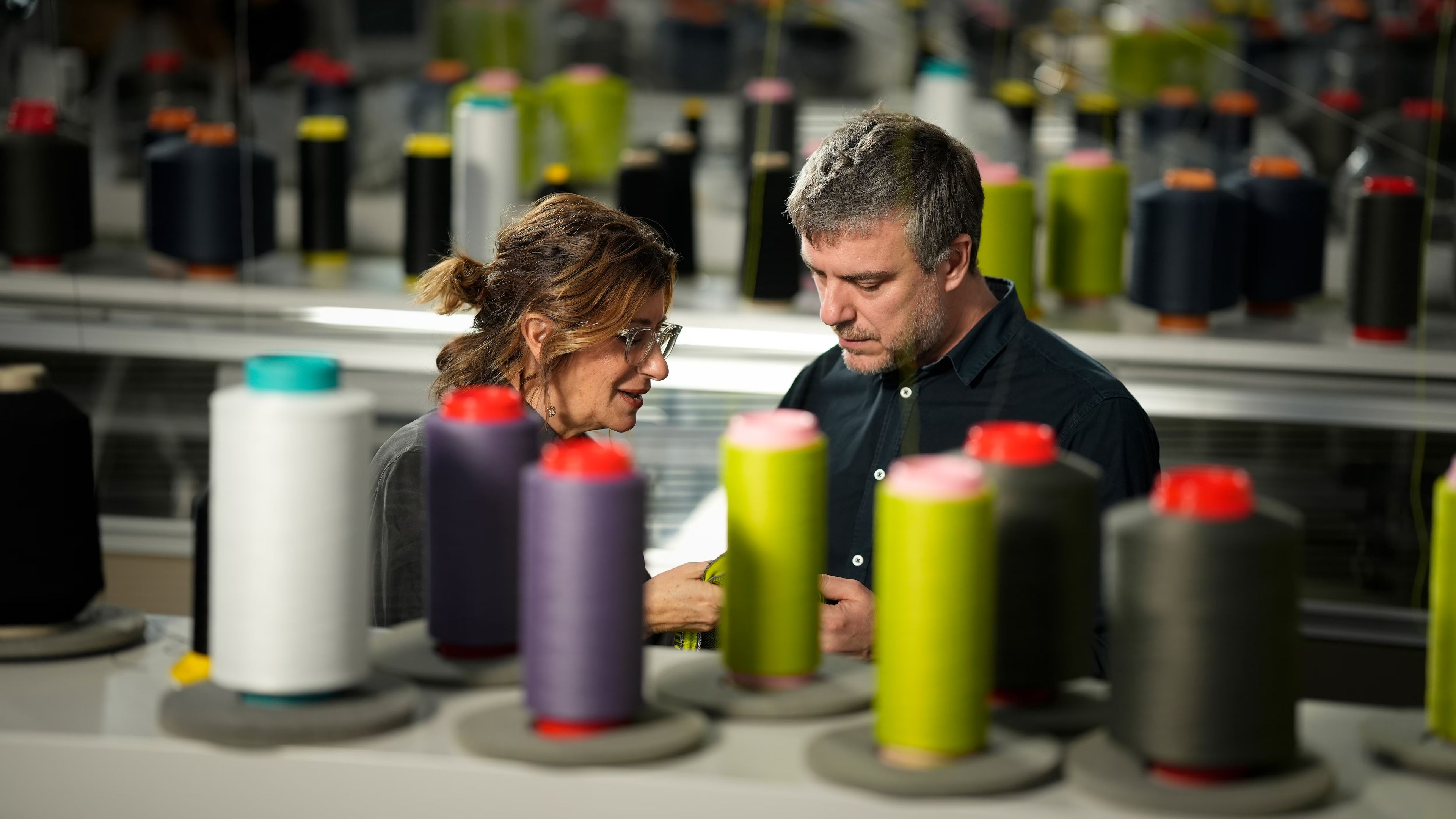 Dve osebi v proizvodnem prostoru pregledujeta tekstilni material med številnimi tulci niti različnih barv. V ospredju so razporejeni koluti niti, ki nakazujejo proces tekstilne izdelave.