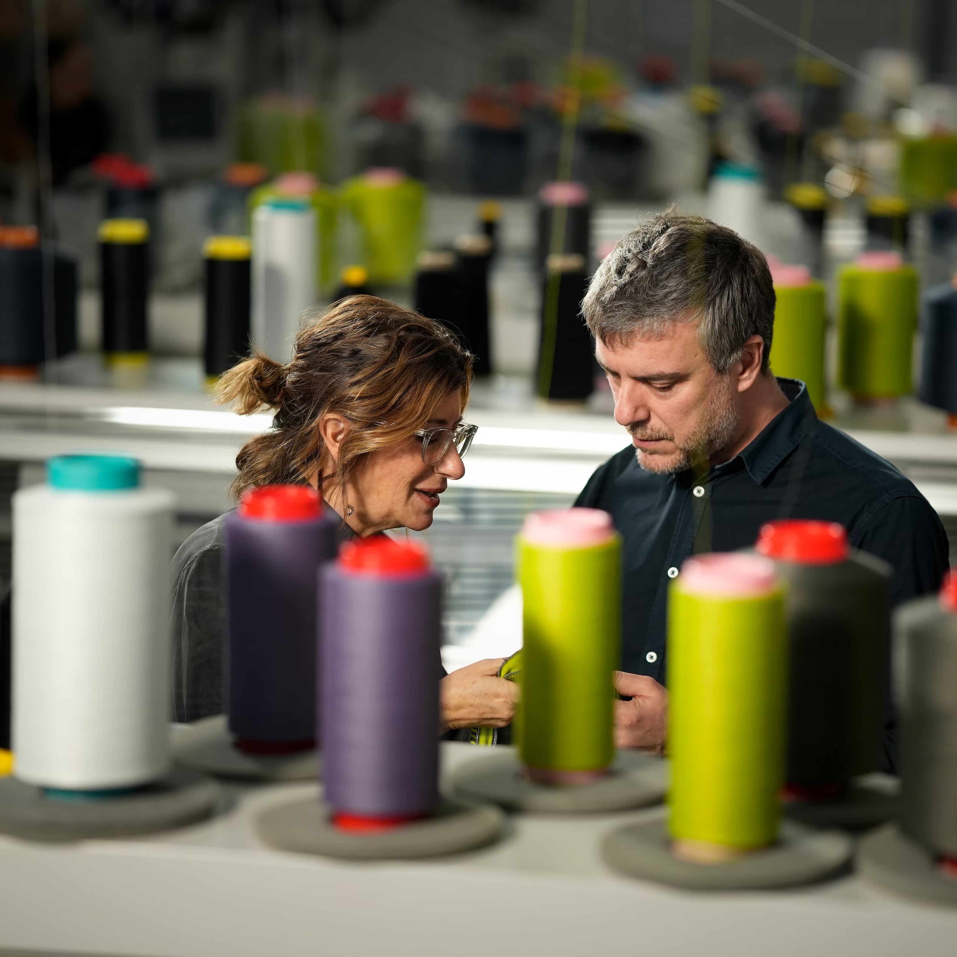 Dve osebi v proizvodnem prostoru pregledujeta tekstilni material med številnimi tulci niti različnih barv. V ospredju so razporejeni koluti niti, ki nakazujejo proces tekstilne izdelave.
