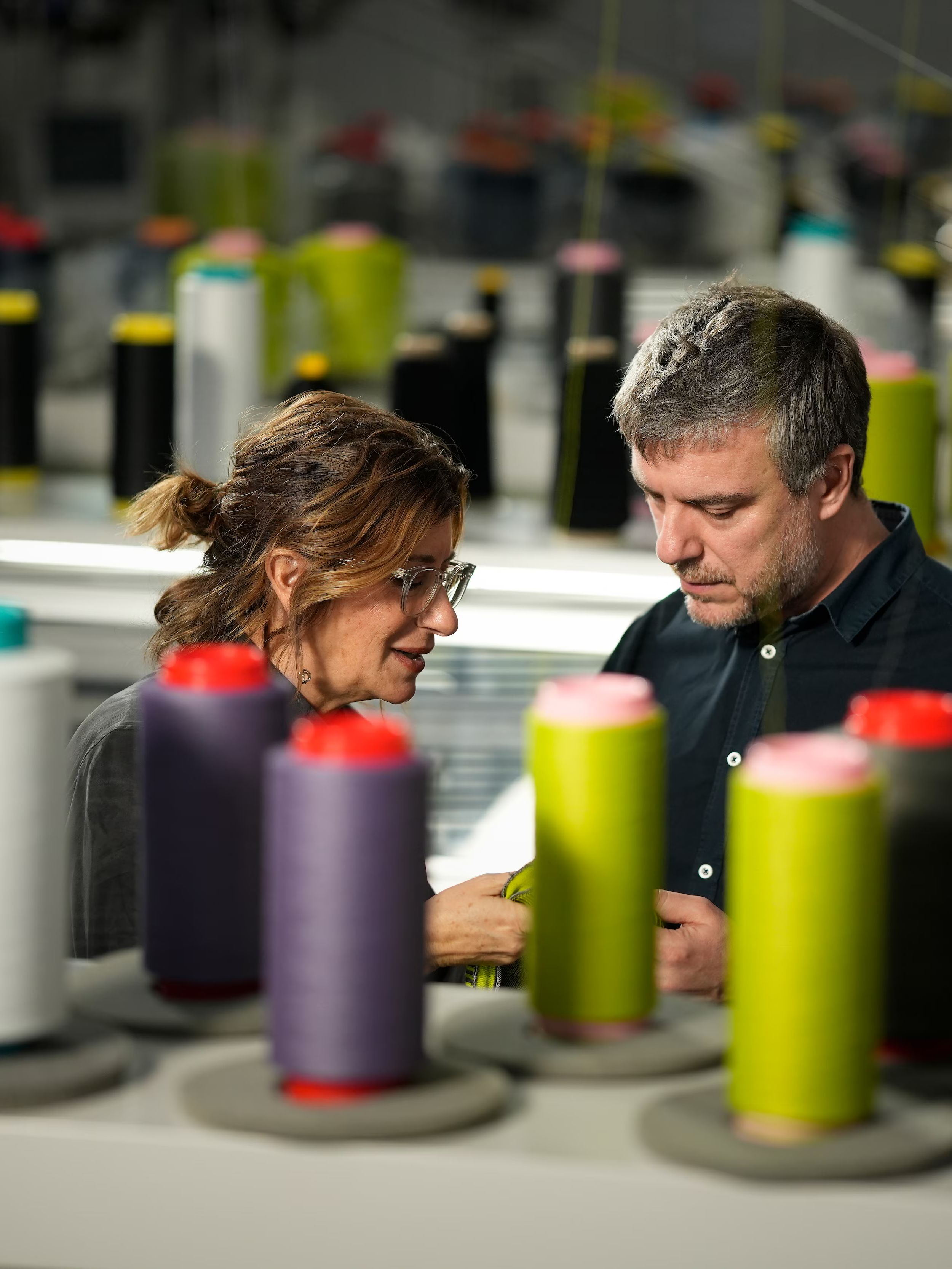 Dve osebi v proizvodnem prostoru pregledujeta tekstilni material med številnimi tulci niti različnih barv. V ospredju so razporejeni koluti niti, ki nakazujejo proces tekstilne izdelave.