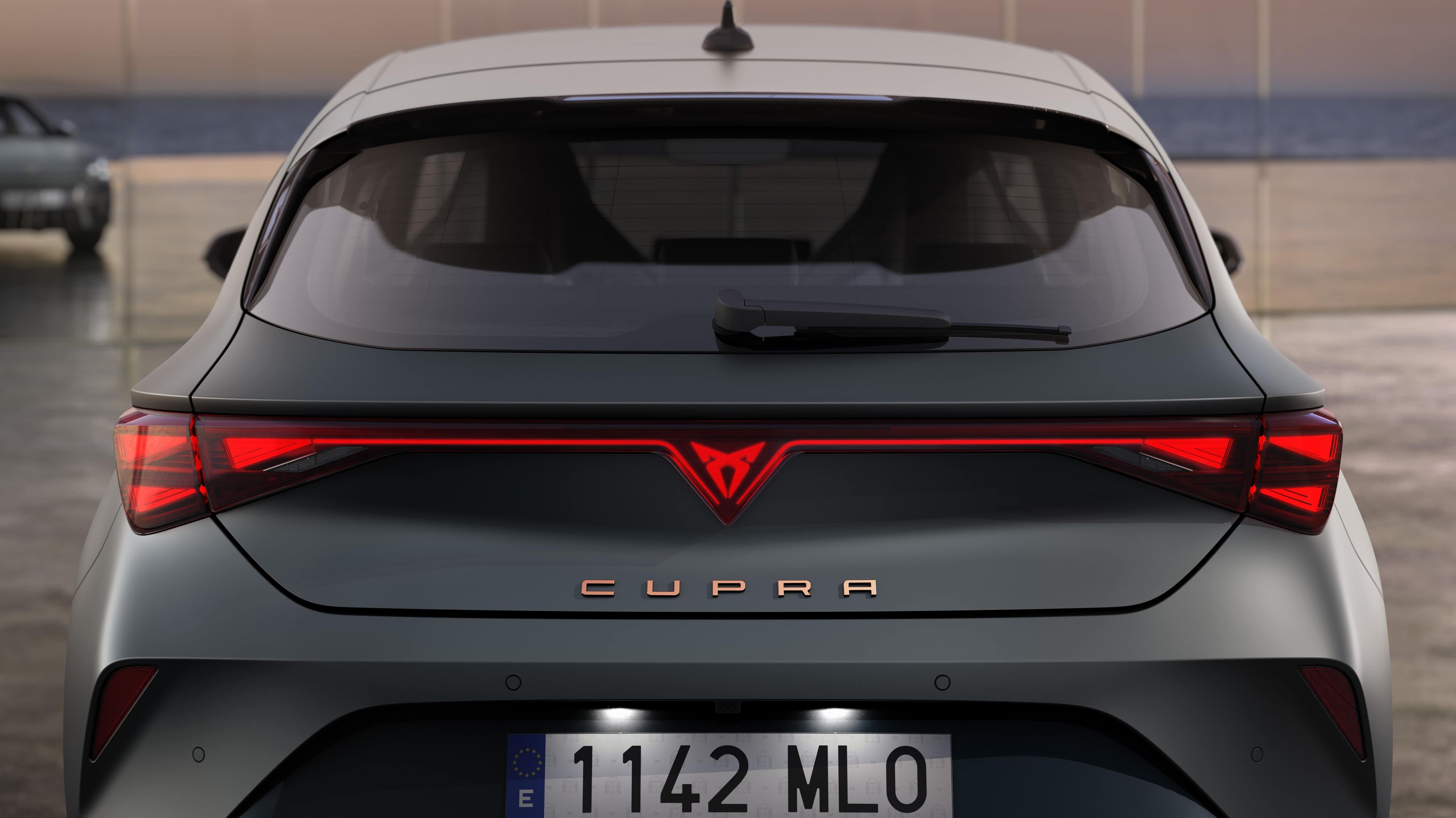 Zadnji del sivega avtomobila znamke Cupra Leon, parkiranega pred steklenimi vrati, z vidno registrsko tablico '1142 MLO'. V ozadju je še en avtomobil, morje in nejasno nebo.