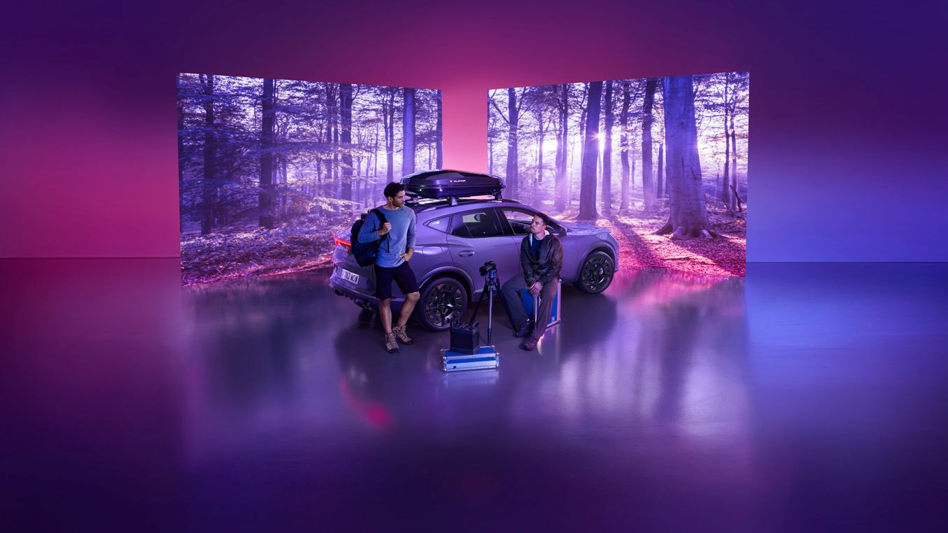 Na sliki sta dva človeka, ki stojita pred avtomobilom v digitalni postavitvi, obdani z zaslonom, ki prikazuje gozdno pokrajino. Na tleh pred avtomobilom so kovčki in športna oprema. Ozadje je vijolično osvetljeno.
