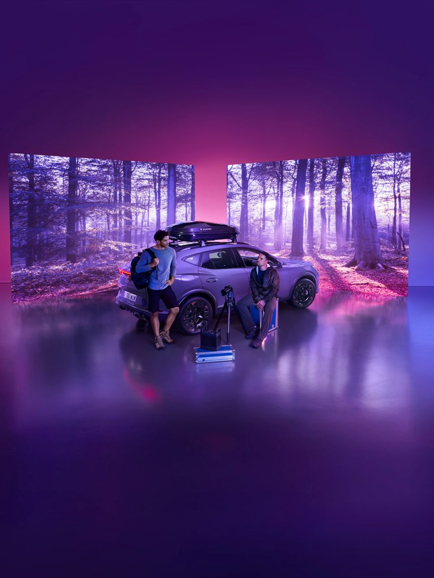 Na sliki sta dva človeka, ki stojita pred avtomobilom v digitalni postavitvi, obdani z zaslonom, ki prikazuje gozdno pokrajino. Na tleh pred avtomobilom so kovčki in športna oprema. Ozadje je vijolično osvetljeno.