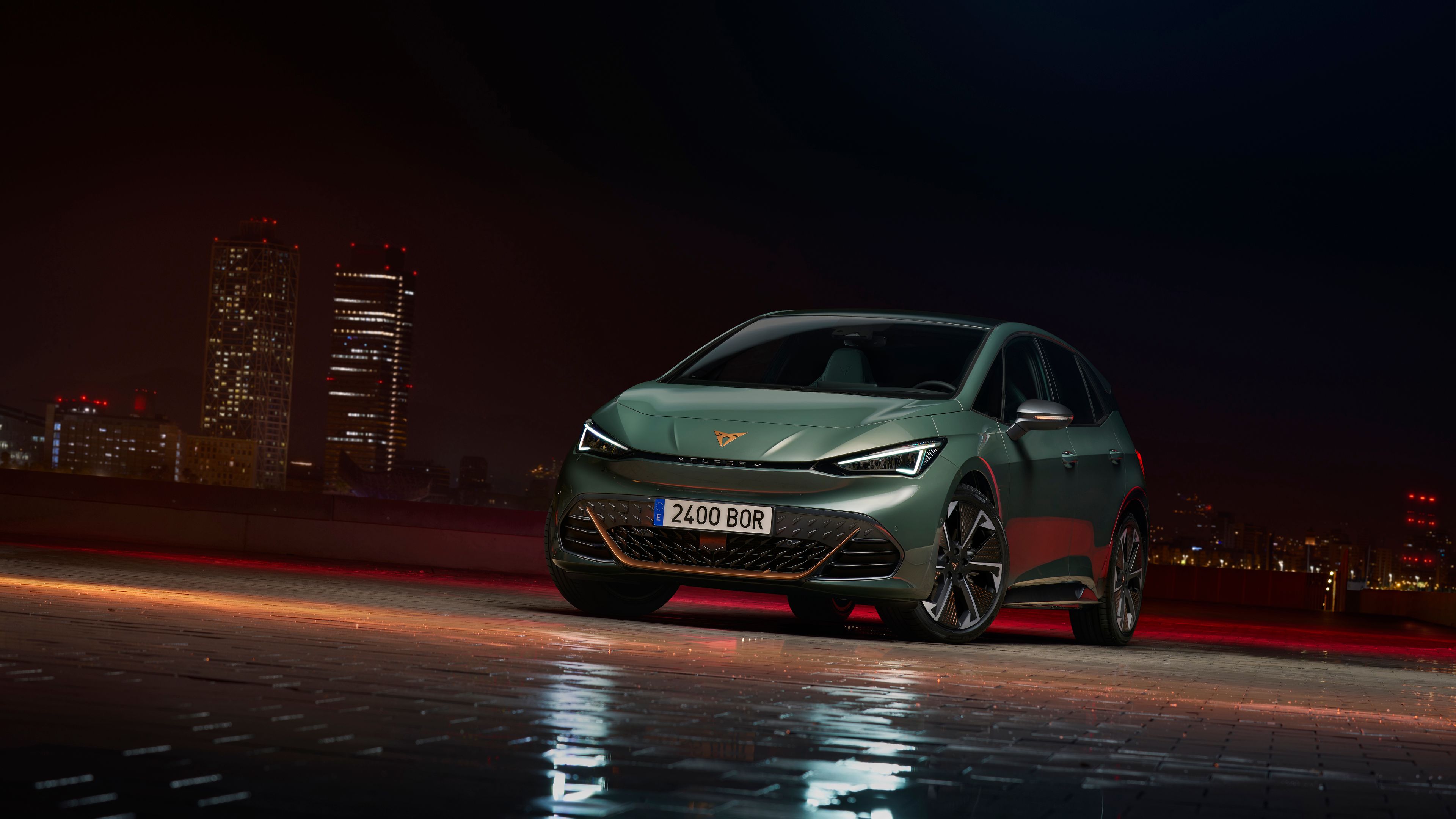 Temno zelen CUPRA Born stoji na mokrem tlakovcu v mestnem okolju v nočnem času. V ozadju so vidne osvetljene stolpnice. Avto ima moderna aluminijasta platišča.