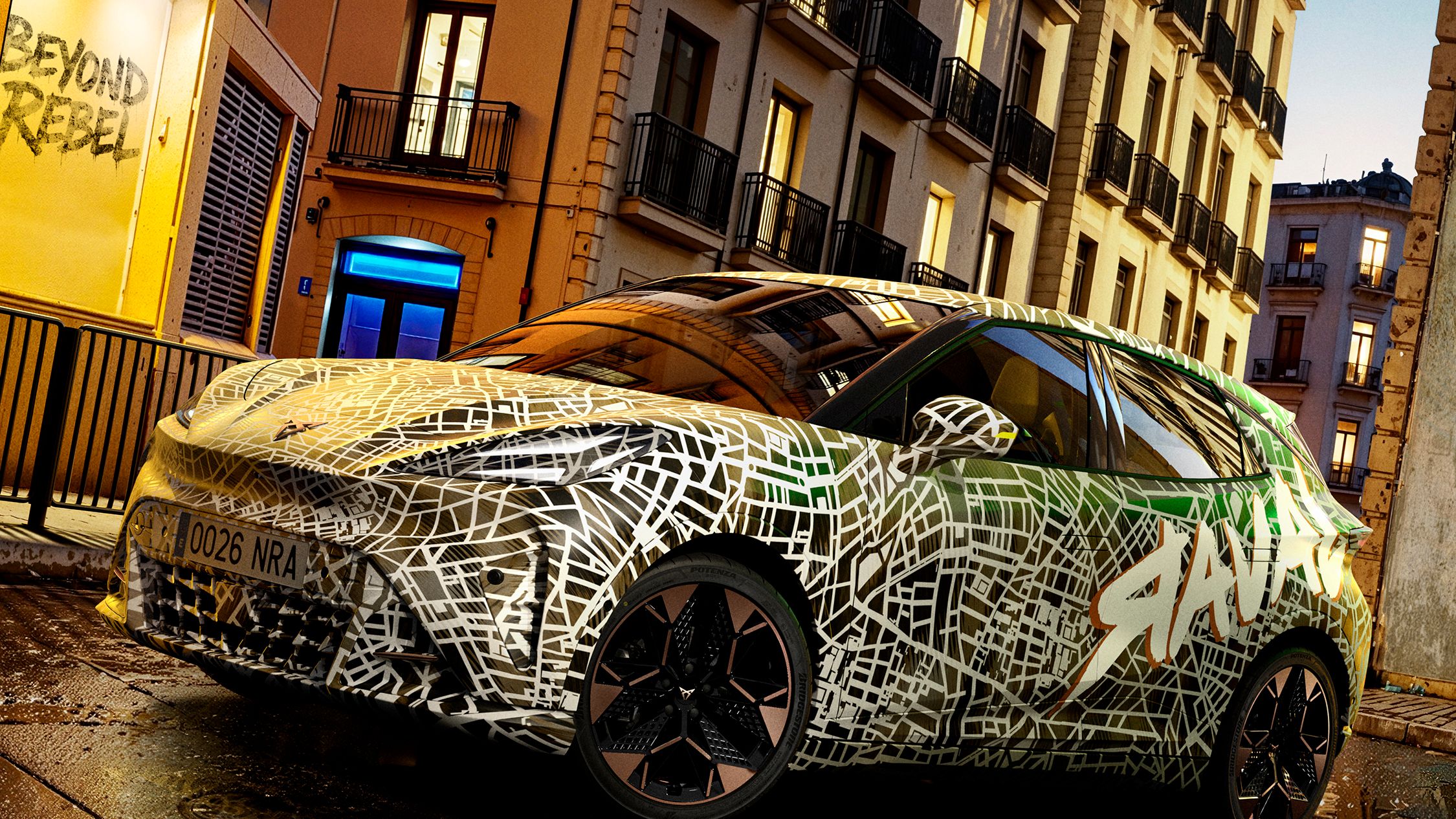 Avtomobil Cupra Raval z grafičnimi vzorci parkiran na mokri ulici ob stolpnici. Ulica je slikana ponoči, z osvetljenimi okni na stavbah v ozadju.
