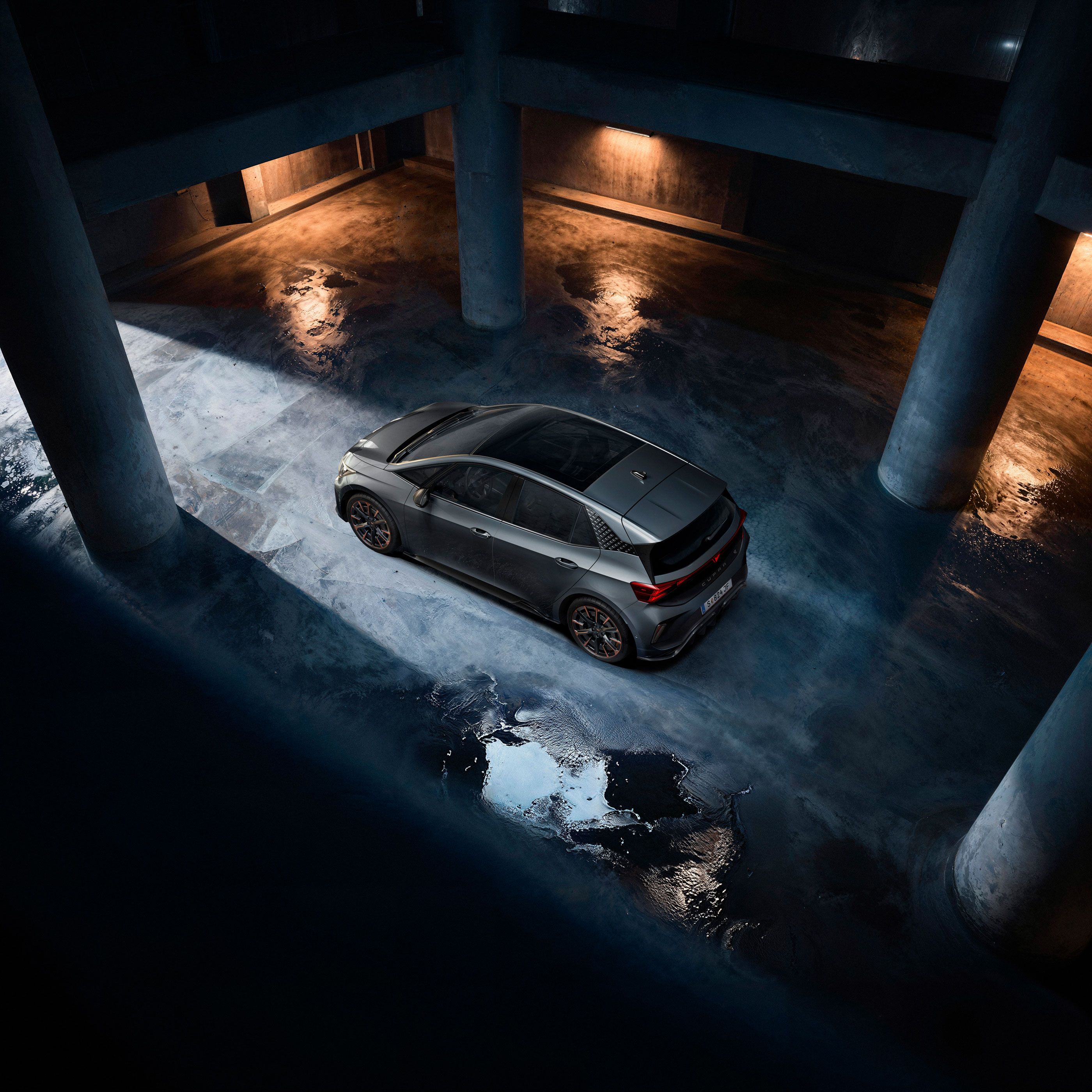 Ein grauer CUPRA Born steht in einer dunklen Tiefgarage. Das Bild ist aus der Vogelperspektive aufgenommen und zeigt Lichterscheinungen auf dem nassen Betonboden. Die Szenerie ist durch indirektes Licht beleuchtet, das eine dramatische Atmosphäre schafft.