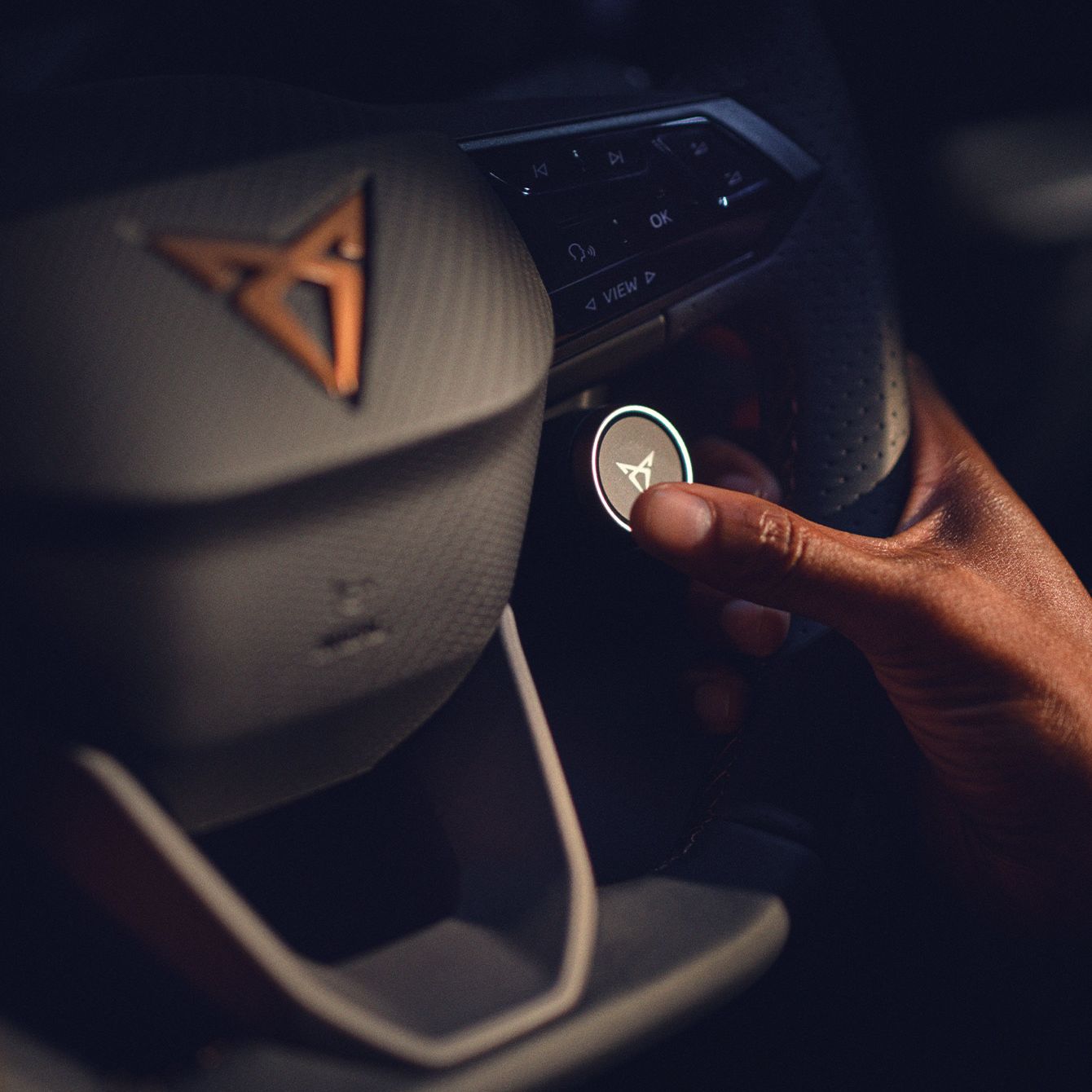 Oseba pritiska na osvetljeno stikalo na volanu avtomobila CUPRA Born. Na volanu je vidna značka avtomobilske znamke Cupra. 