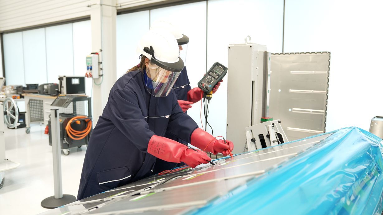 Tehnik v zaščitni opremi (čelada, vizir in izolacijske rokavice) v laboratoriju z multimetrom preverja električne povezave na velikem baterijskem modulu.