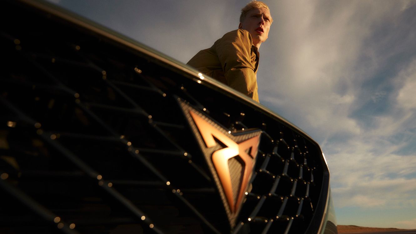 Moški stoji za avtomobilom CUPRA Formentor na katerem se jasno vidi logo znamke. Ozadje prikazuje modro-sive oblake na nebu.