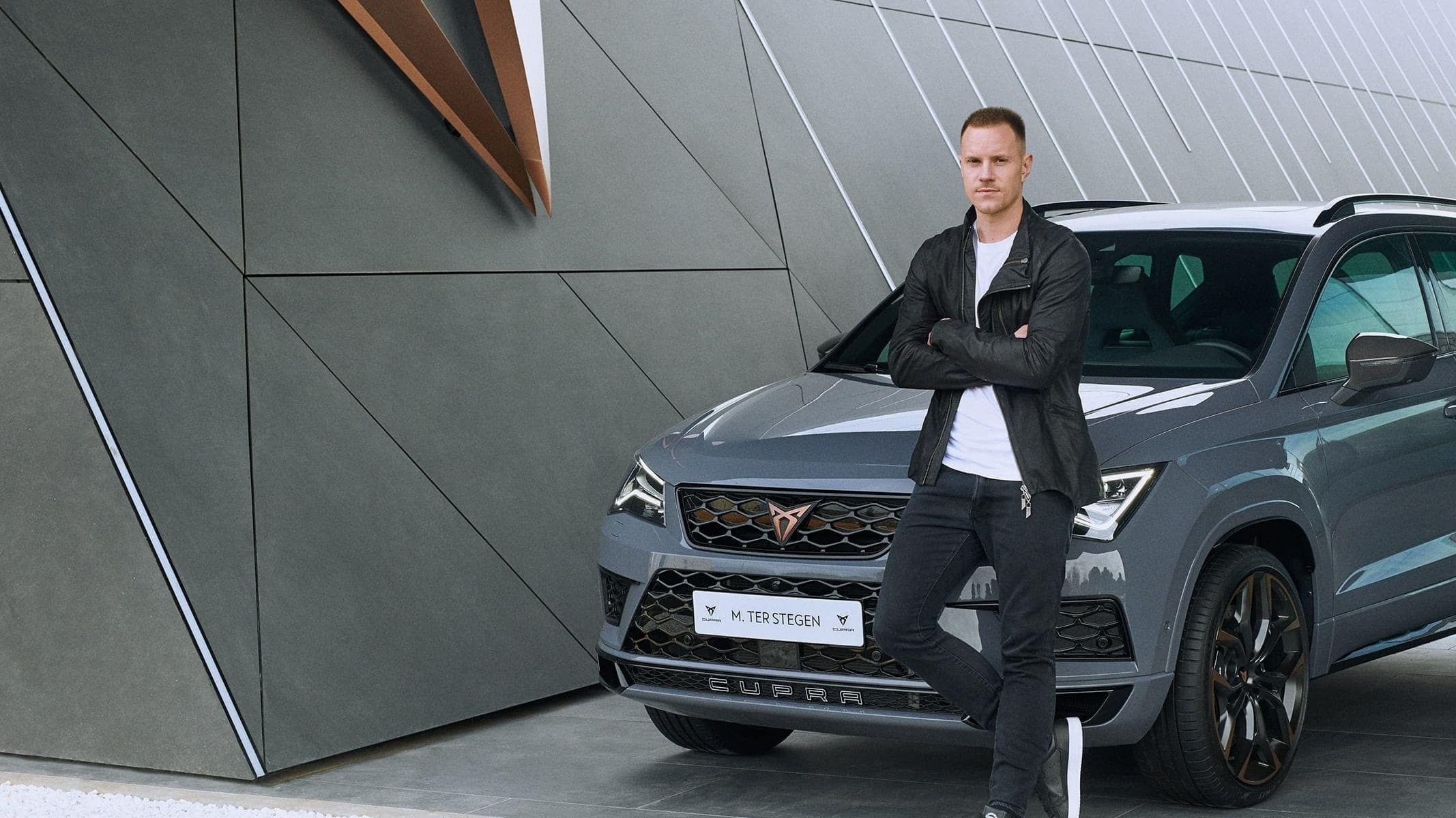Marc ter Stegen, ki je postal uradni ambasador znamke CUPRA stoji pred CUPRA vozilom. Vozilo je parkirano pred moderno arhitekturno strukturo.