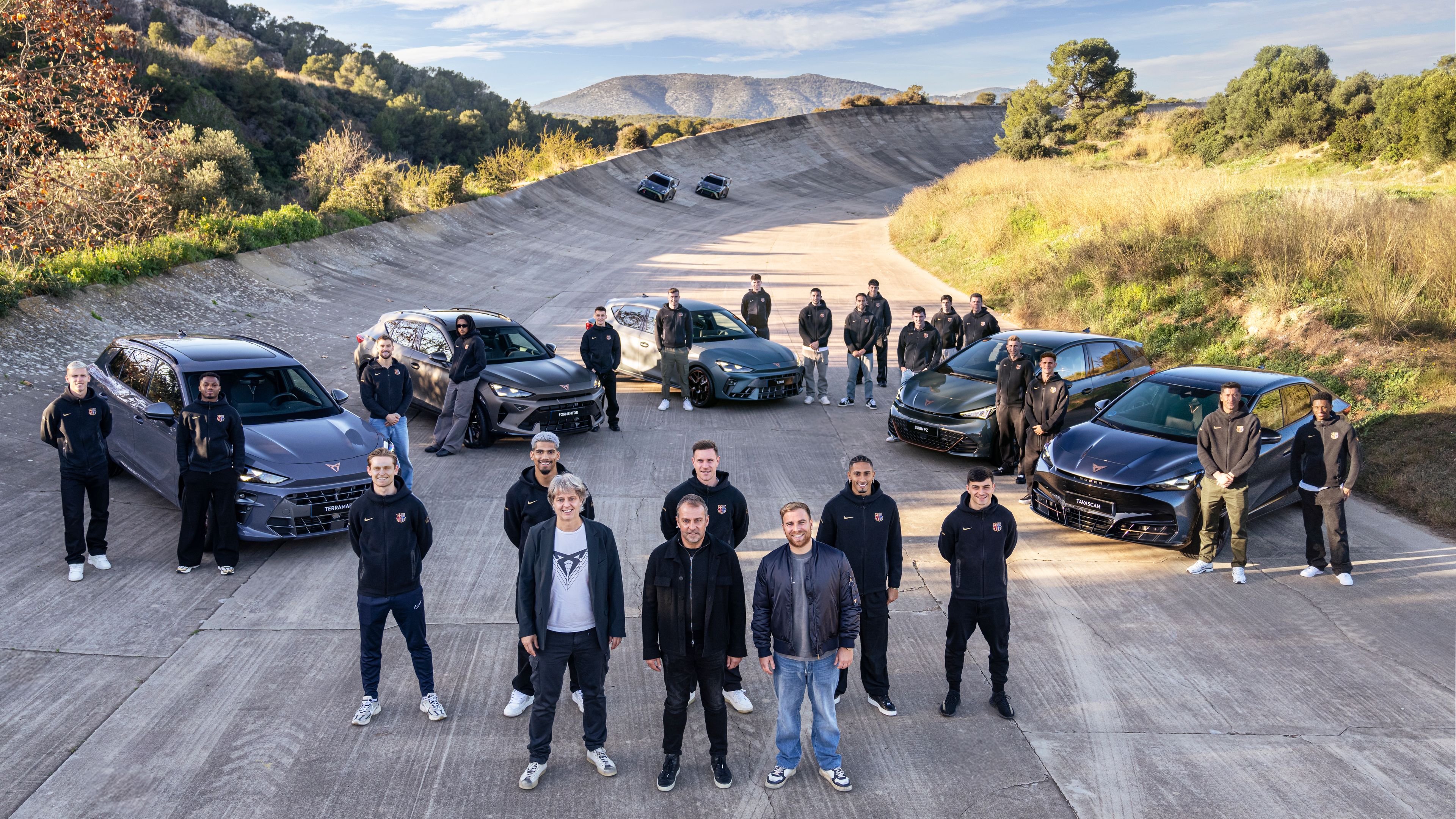 Skupina ljudi stoji na dirkališču ob več športnih vozilih znamke CUPRA v dinamični formaciji. Prizor poudarja ekipni duh in zmogljivost avtomobilov v edinstvenem, odprtem okolju.