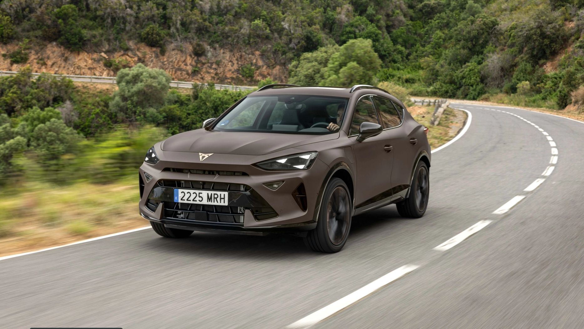 Na posnetku je moderno rjavo vozilo znamke CUPRA, ki vozi po ovinkasti cesti obdan z bujno vegetacijo. Sprednji del avtomobila je dinamičnega videza.