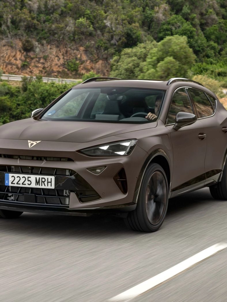 Na posnetku je moderno rjavo vozilo znamke CUPRA, ki vozi po ovinkasti cesti obdan z bujno vegetacijo. Sprednji del avtomobila je dinamičnega videza.