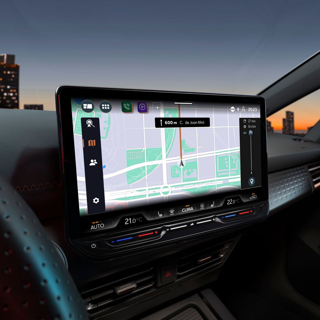 Notranjost avtomobila Cupra Born prikazuje osrednjo konzolo z velikim digitalnim zaslonom, ki prikazuje zemljevid in navigacijske ukaze. Poleg navigacije so na zaslonu tudi različne funkcije, kot so nastavitev temperature in predvajanje glasbe. V ozadju je skozi okno vidna mestna panorama ob sončnem zahodu.