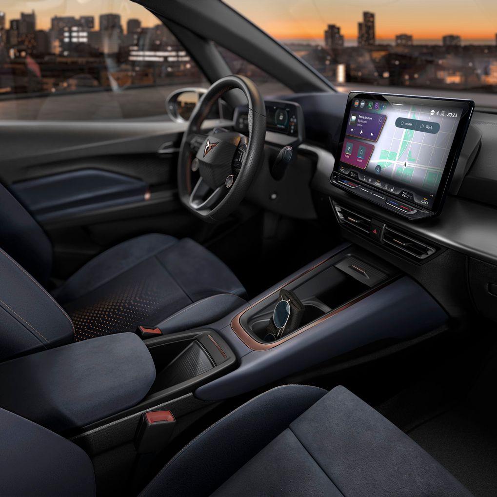 Notranjost sodobnega vozila CUPRA Born z velikim zaslonom infotainment sistema, volanom in upravljalnimi elementi. Vozilo ima elegantne črne sedeže, skozi okna pa se vidi sončni zahod nad osvetljenim mestom.