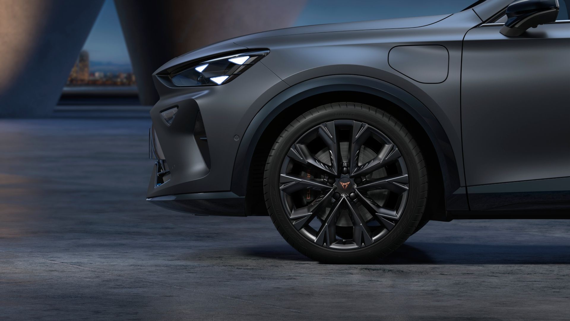 Sprednji del sodobnega avtomobila CUPRA Formentpor PA v temno sivi mat barvi, parkiranega na betonski površini ob mraku. Na sliki so vidne elegantne luči in sprednji del avtomobilskega platišča. V ozadju se nahaja osvetljen mestni pejsaž.