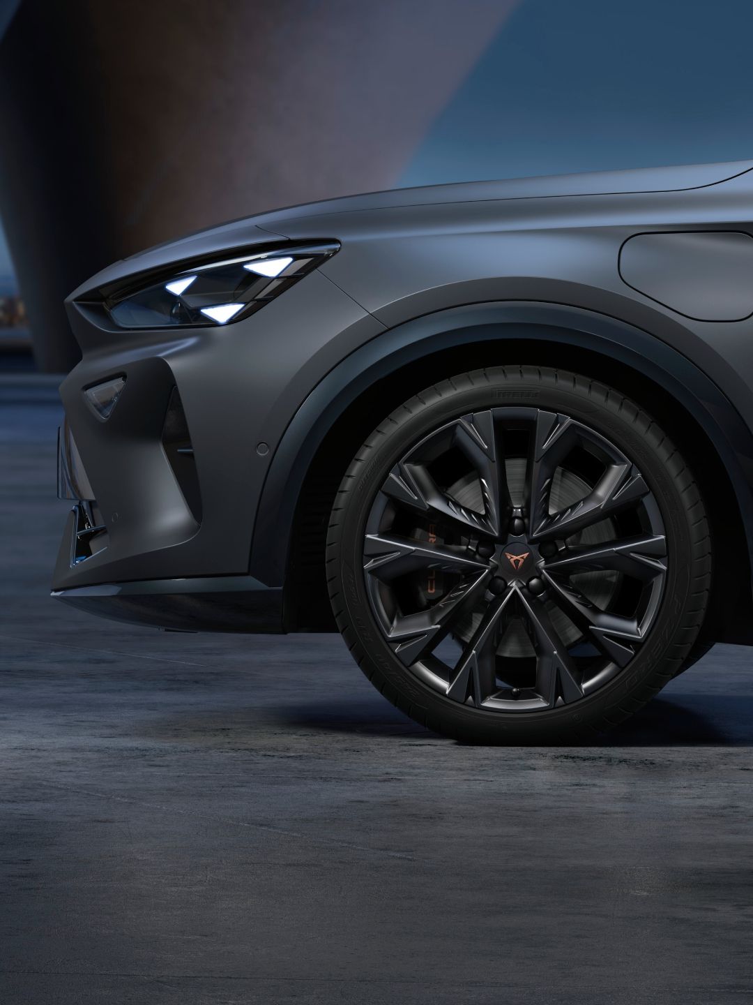 Sprednji del sodobnega avtomobila CUPRA Formentpor PA v temno sivi mat barvi, parkiranega na betonski površini ob mraku. Na sliki so vidne elegantne luči in sprednji del avtomobilskega platišča. V ozadju se nahaja osvetljen mestni pejsaž.