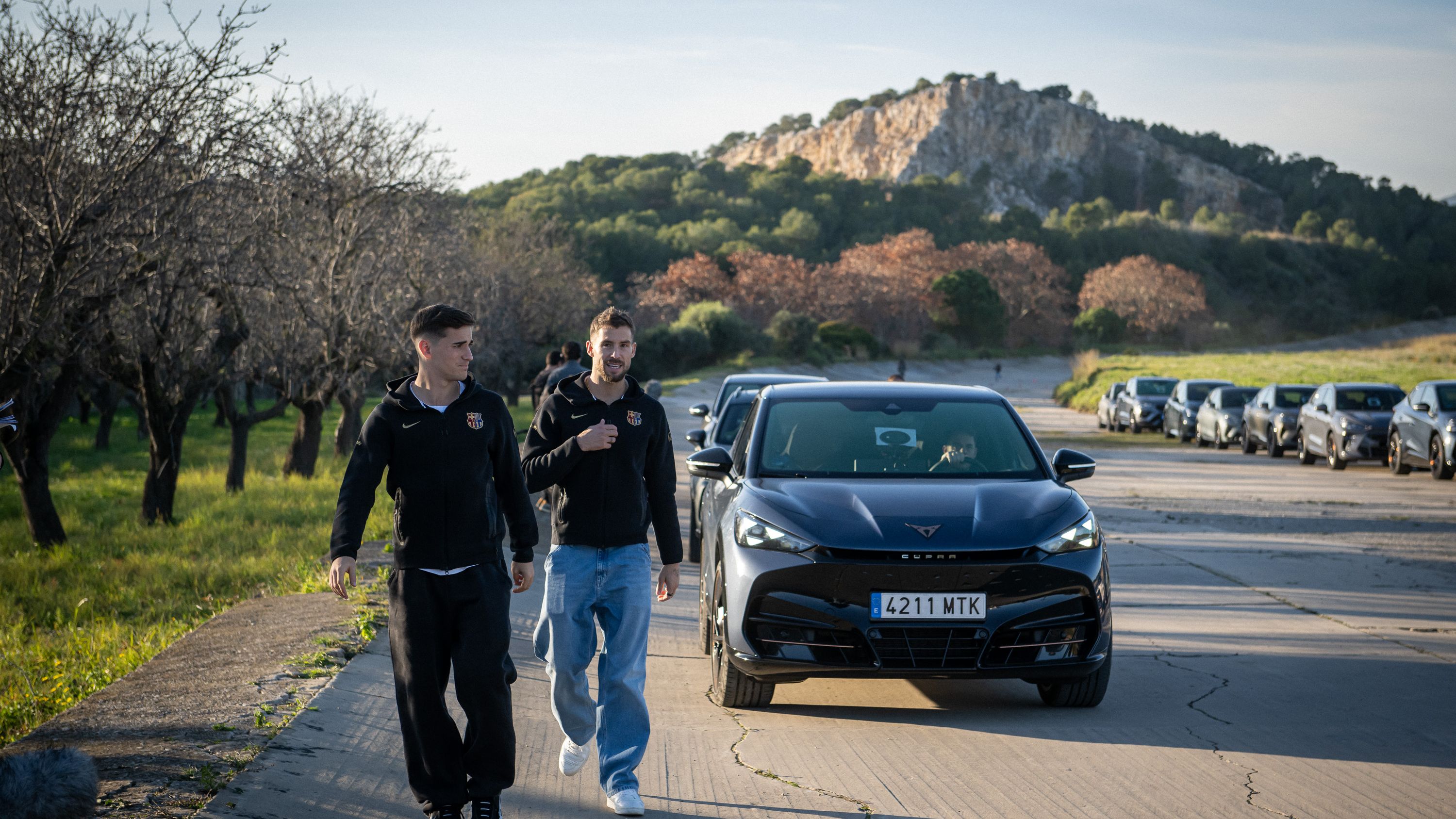 Dva igralca FC Barcelona hodita po asfaltni poti ob parkiranem črnem avtomobilu CUPRA Terramar. V ozadju so drevesa in skalnata vzpetina pod jasnim nebom. Na desni strani je vrsta parkiranih avtomobilov.