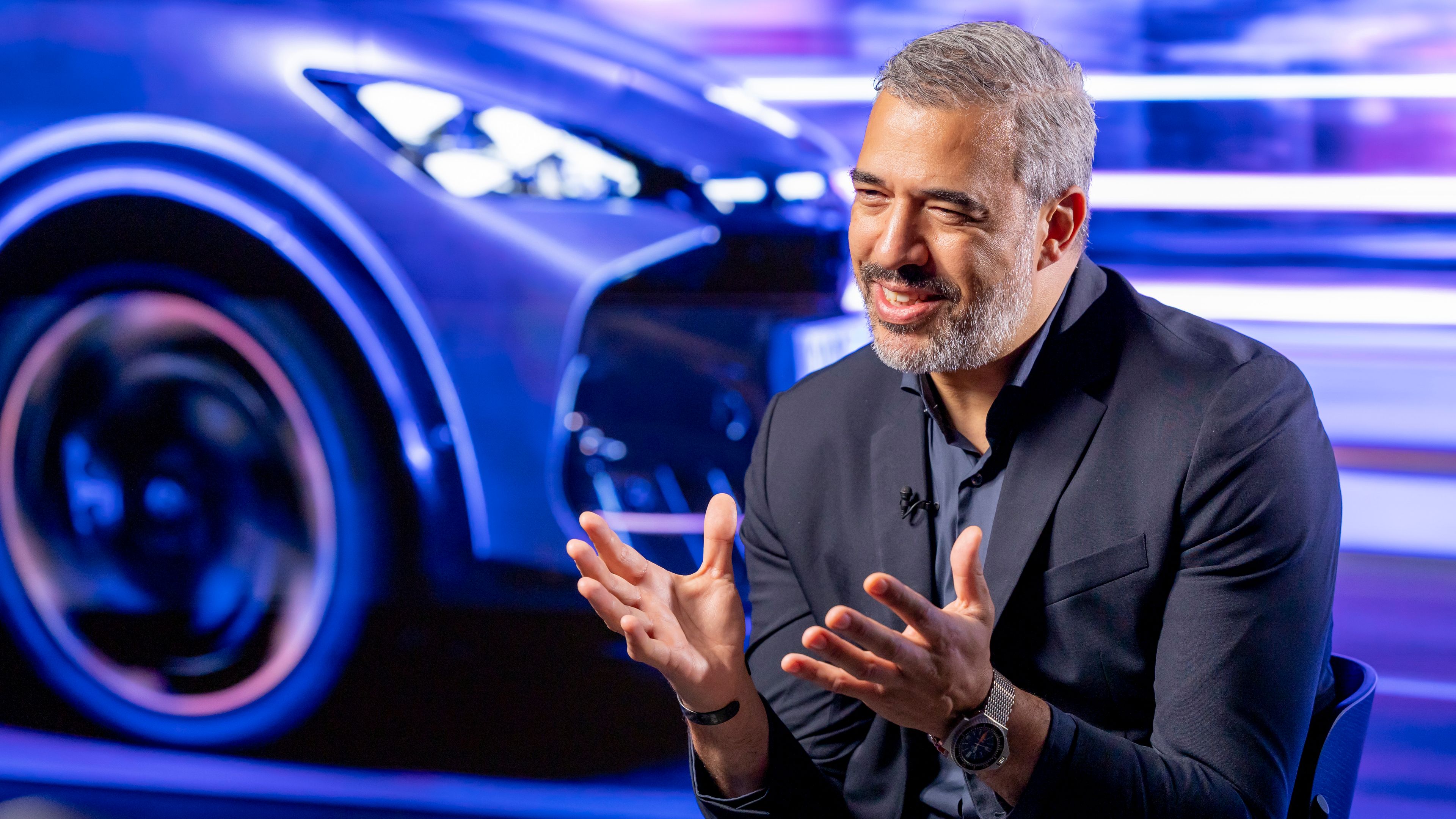 Oblikovalec  znamke CUPRA Jorge Díez, v elegantni temni obleki sedi pred ozadjem, ki prikazuje del avtomobila. Oseba ima sive lase in razložene roke, kot da nekaj pojasnjuje. Ozadje je živahno in barvito.