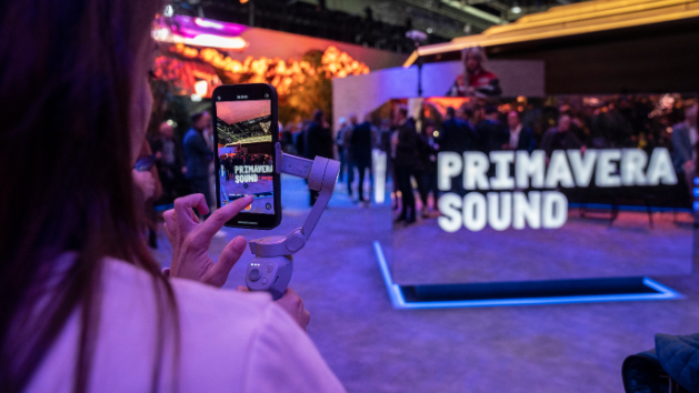 Oseba drži telefon, s katerim snema dogodek. V ozadju je množica ljudi in osvetljen napis "Primavera Sound". Okolica je živahna in polna barvnih luči.