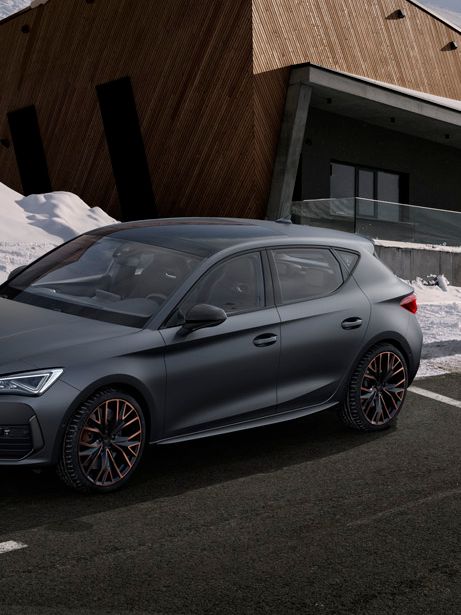 CUPRA Leon 5D je parkiran na asfaltiranem parkirišču pred sodobno zgradbo, ki stoji v zasneženi pokrajini. V bližini avtomobila sta dve osebi – ženska stoji, moški pa hodi proti avtomobilu. V ozadju so zasnežene gore in oblačno nebo.