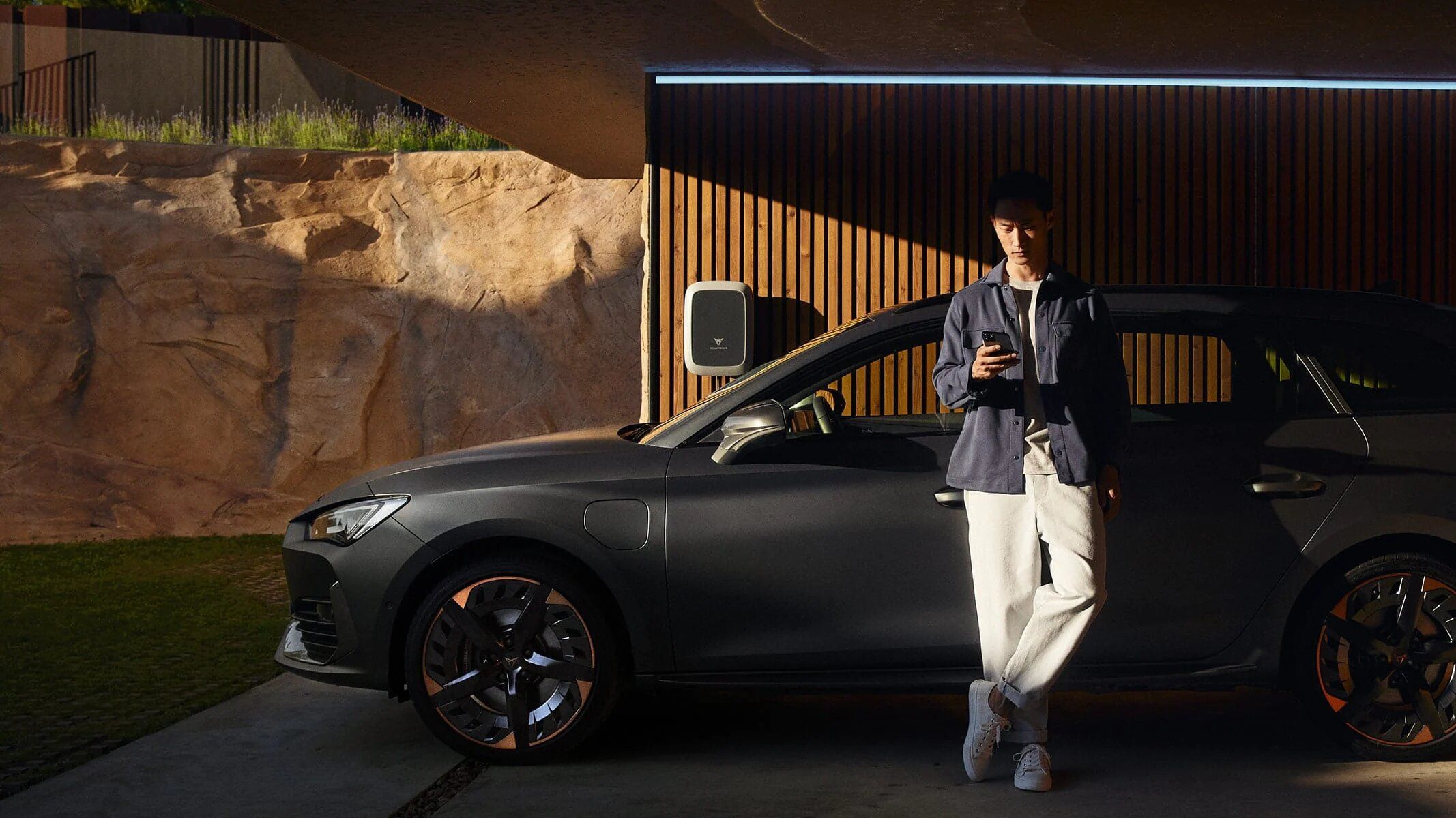 Moški stoji ob parkiranem temnosivem avtomobilu v sodobni garaži. V roki drži mobilni telefon. 