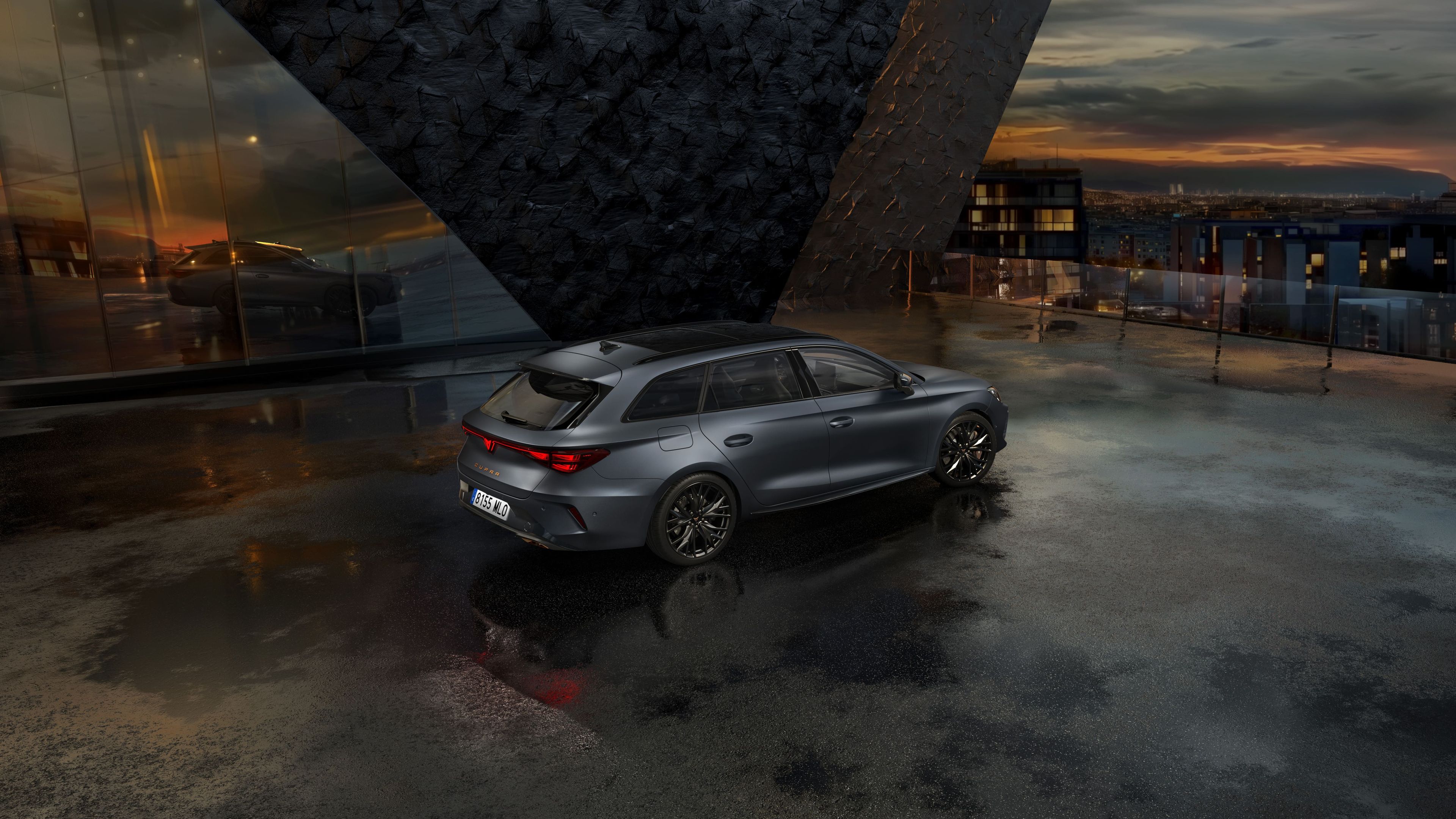 Siva kombilimuzina CUPRA LEON SPORTSTOURER stoji na mokrem asfaltu pred moderno stavbo z velikimi steklenimi okni. Ozadje je oblačno, z odsevom svetlobe na mokri površini. V daljavi se vidijo mestne luči.