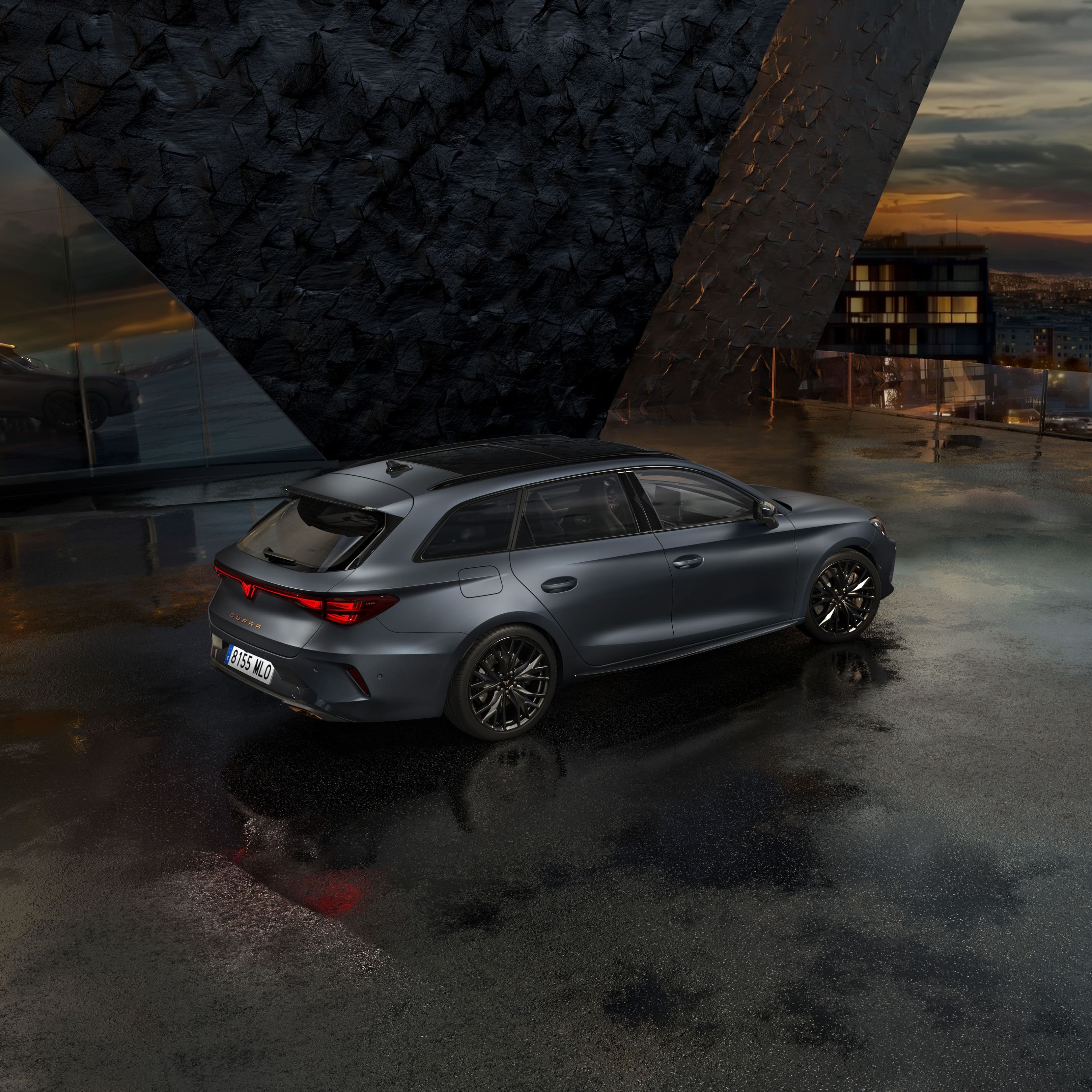 Siva kombilimuzina CUPRA LEON SPORTSTOURER stoji na mokrem asfaltu pred moderno stavbo z velikimi steklenimi okni. Ozadje je oblačno, z odsevom svetlobe na mokri površini. V daljavi se vidijo mestne luči.