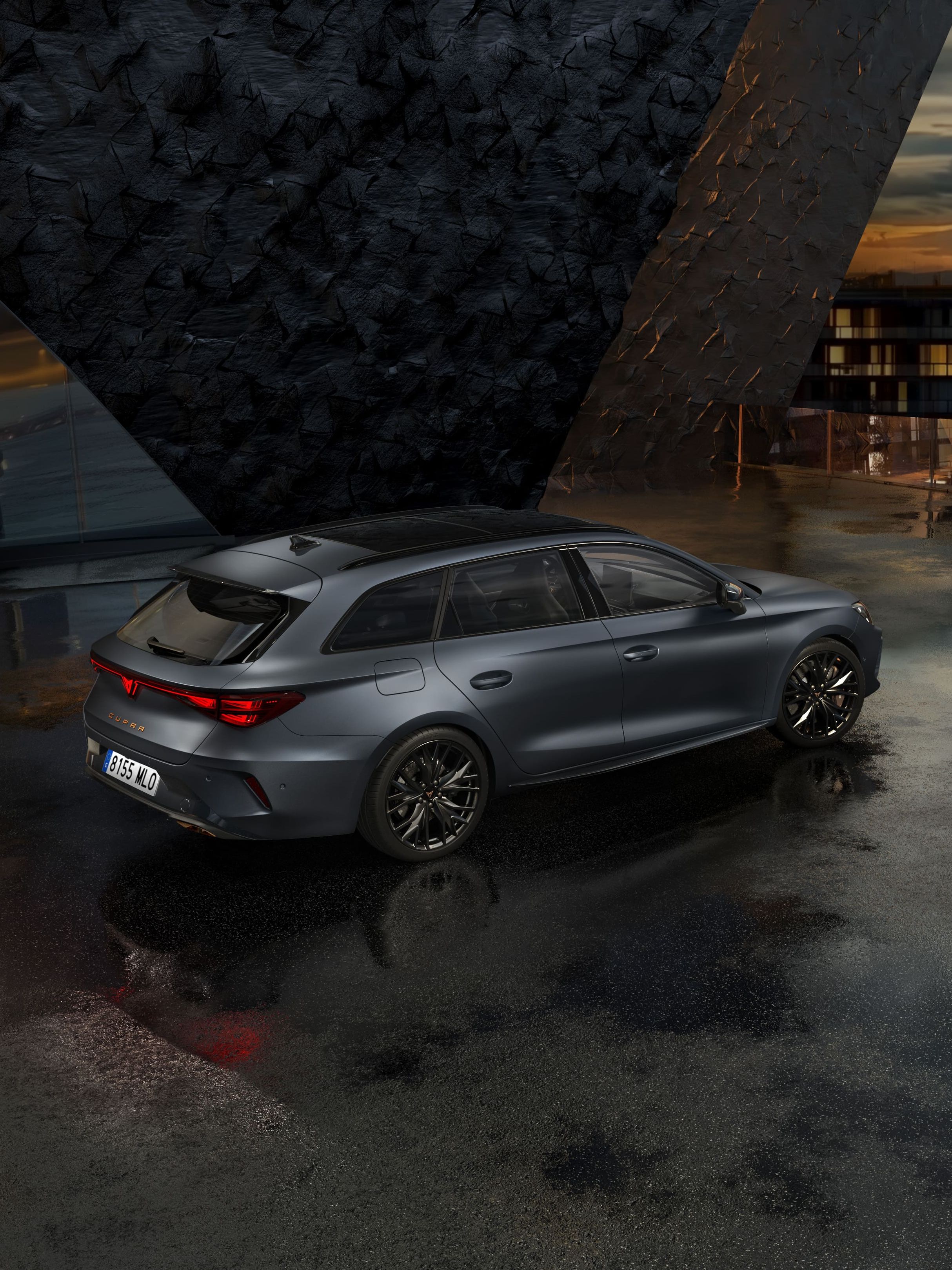 Siva kombilimuzina CUPRA LEON SPORTSTOURER stoji na mokrem asfaltu pred moderno stavbo z velikimi steklenimi okni. Ozadje je oblačno, z odsevom svetlobe na mokri površini. V daljavi se vidijo mestne luči.