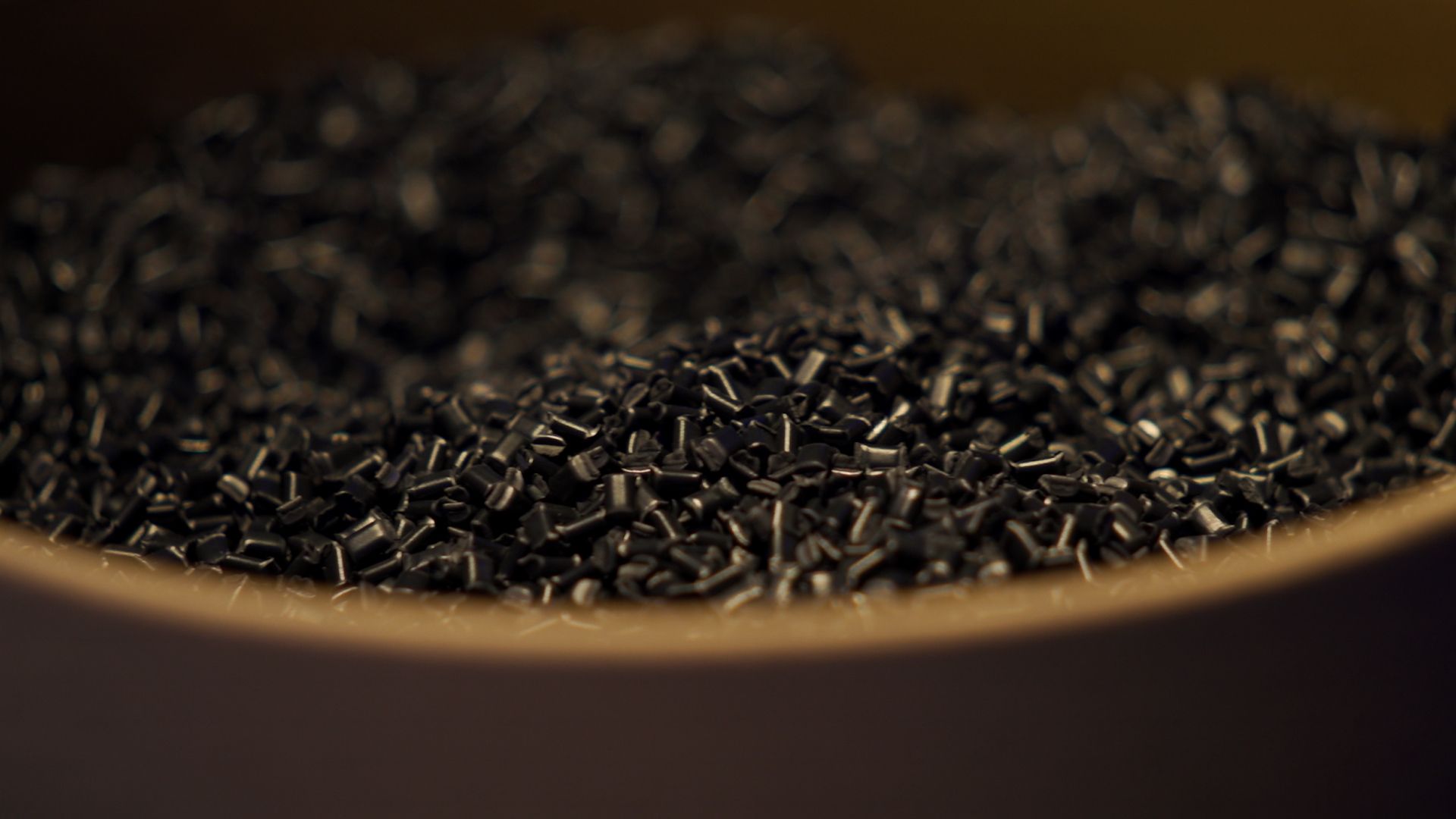 Makro posnetek črnih plastičnih granul v posodi. Majhni valjasti delci so nasuti skupaj, ozadje pa je mehko zamegljeno.