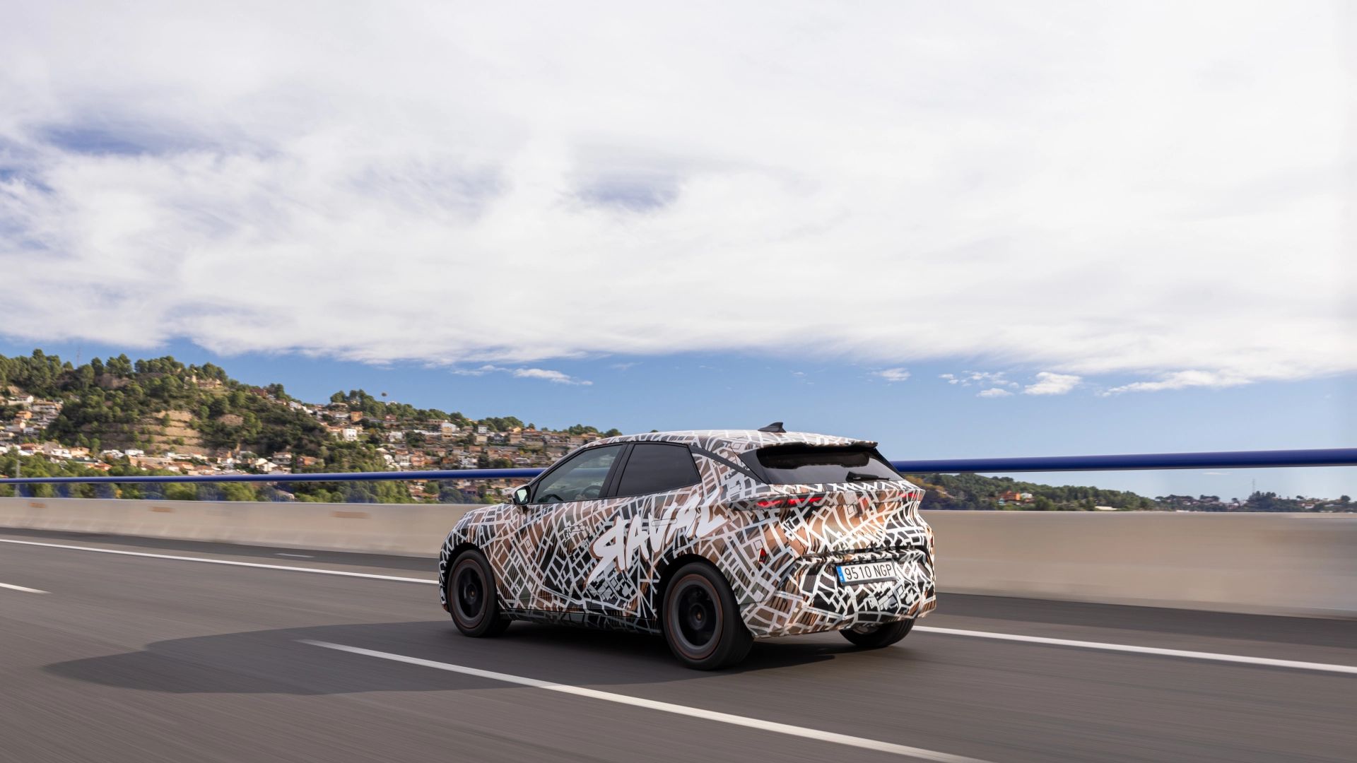 Zamaskiran prototip avtomobila CUPRA Raval Dynamic vozi po mostu, obdan z modro ograjo, v ozadju pa se razprostira zelena pokrajina pod delno oblačnim nebom.